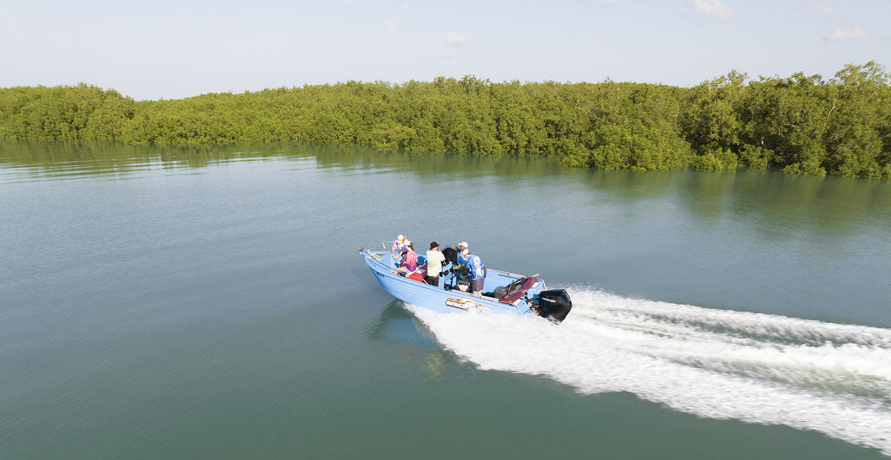 A small blue motorboat with several passengers speeds across calm water, leaving a white wake, with lush green trees in the background.