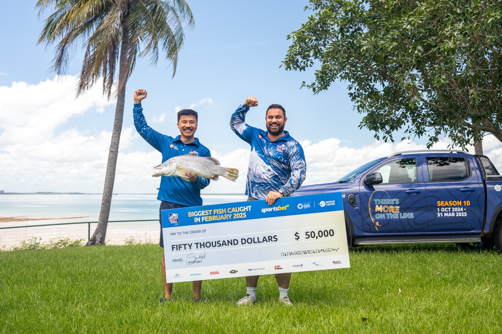 Two men celebrate outdoors with a large fish and a $50,000 prize check. A branded truck is parked nearby under a clear sky.