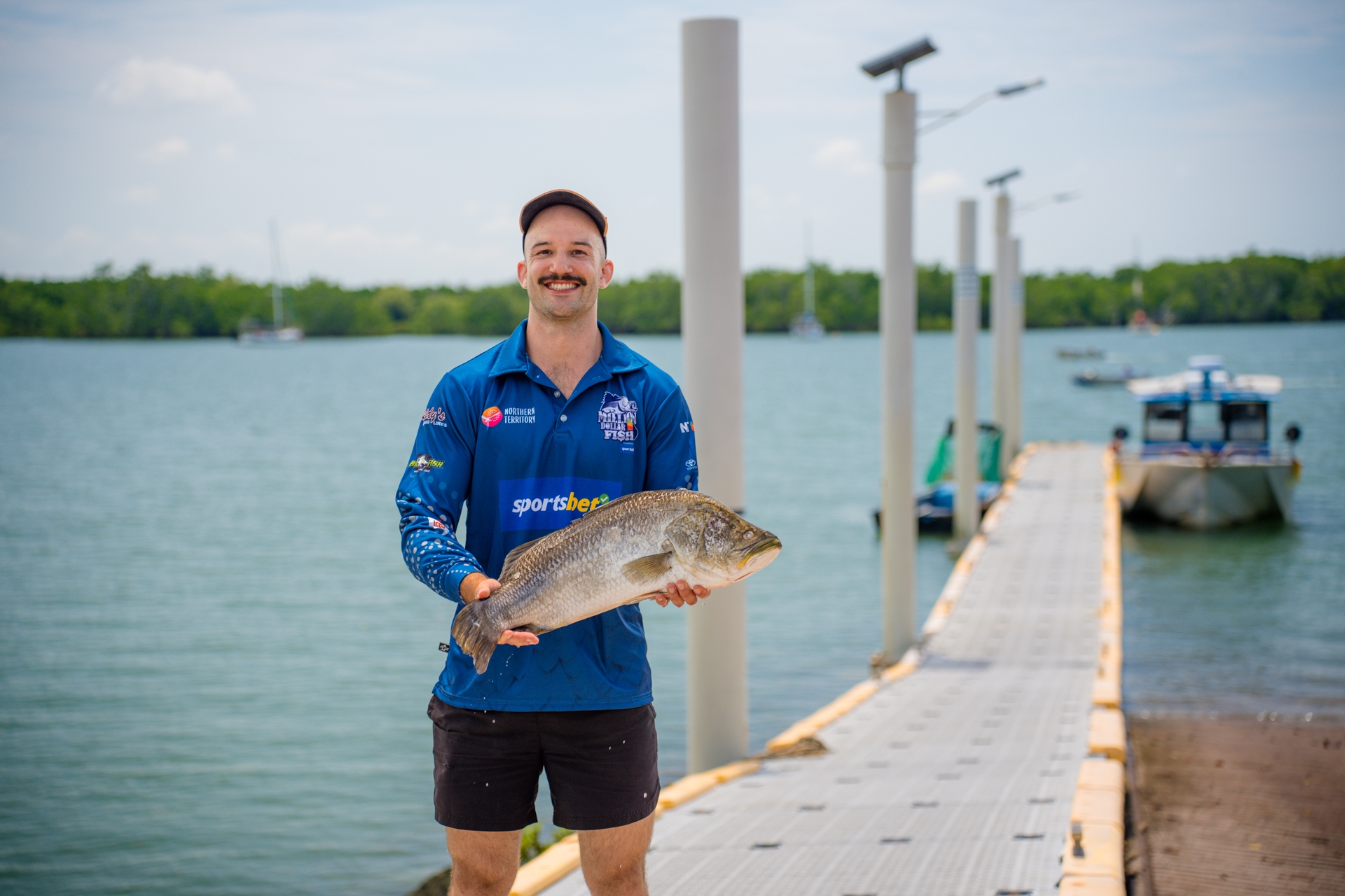 Man holding a large fish on a dock by the water, wearing a blue shirt and cap, with boats and trees in the background.