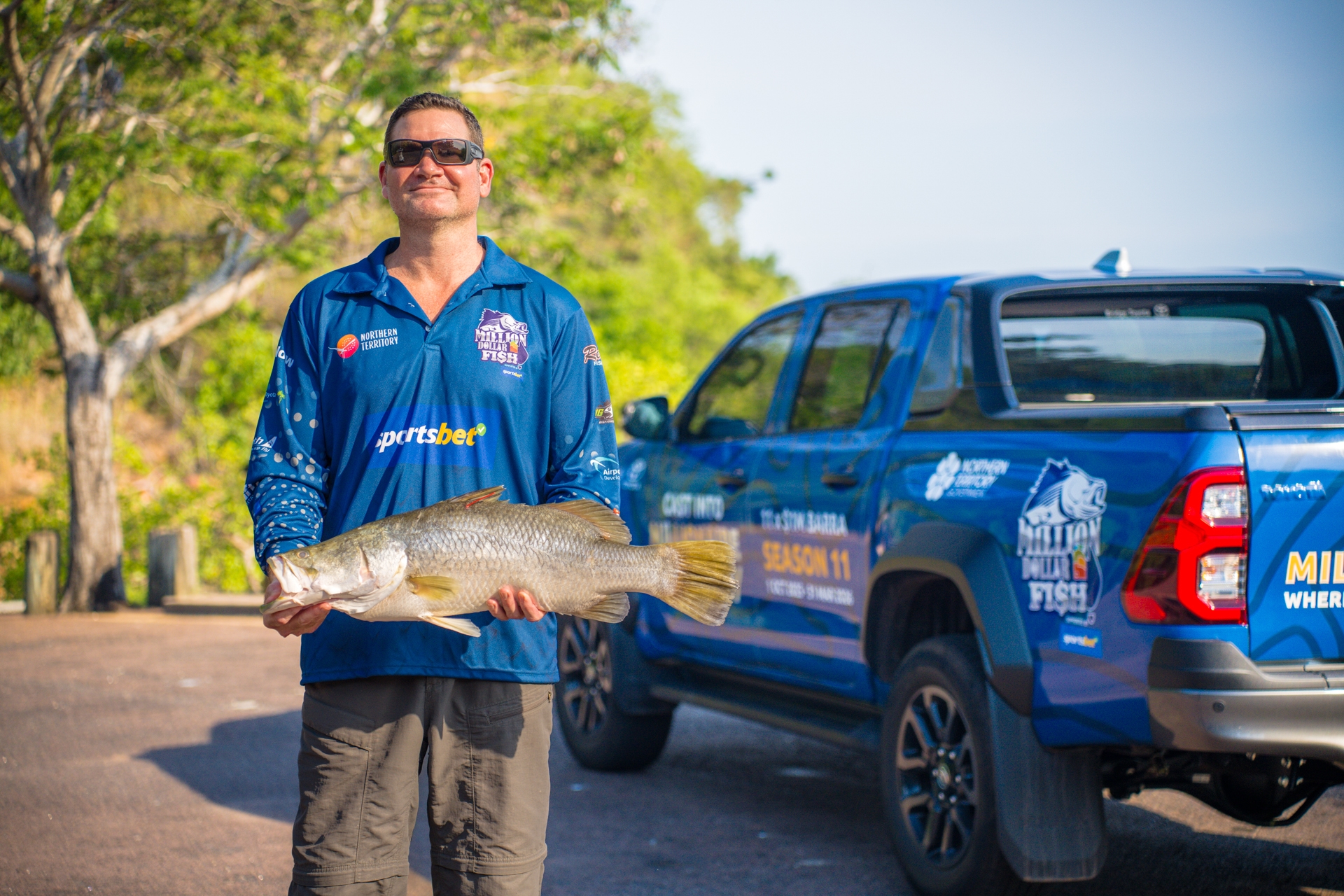 Man holding a large fish stands next to a blue pickup truck with fishing competition branding, surrounded by greenery.