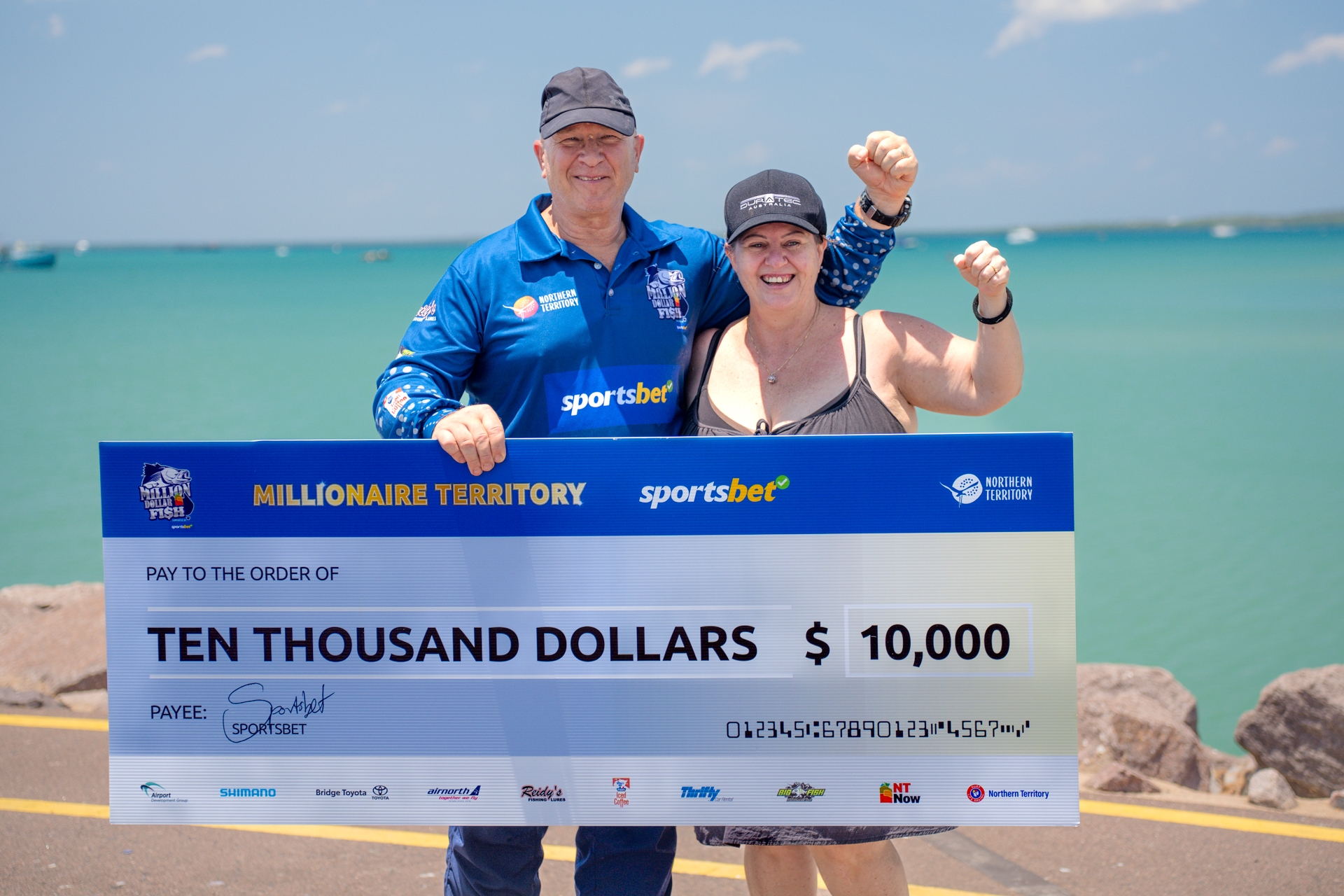 Two smiling people holding a large $10,000 check from Sportsbet, standing outdoors near a body of water.