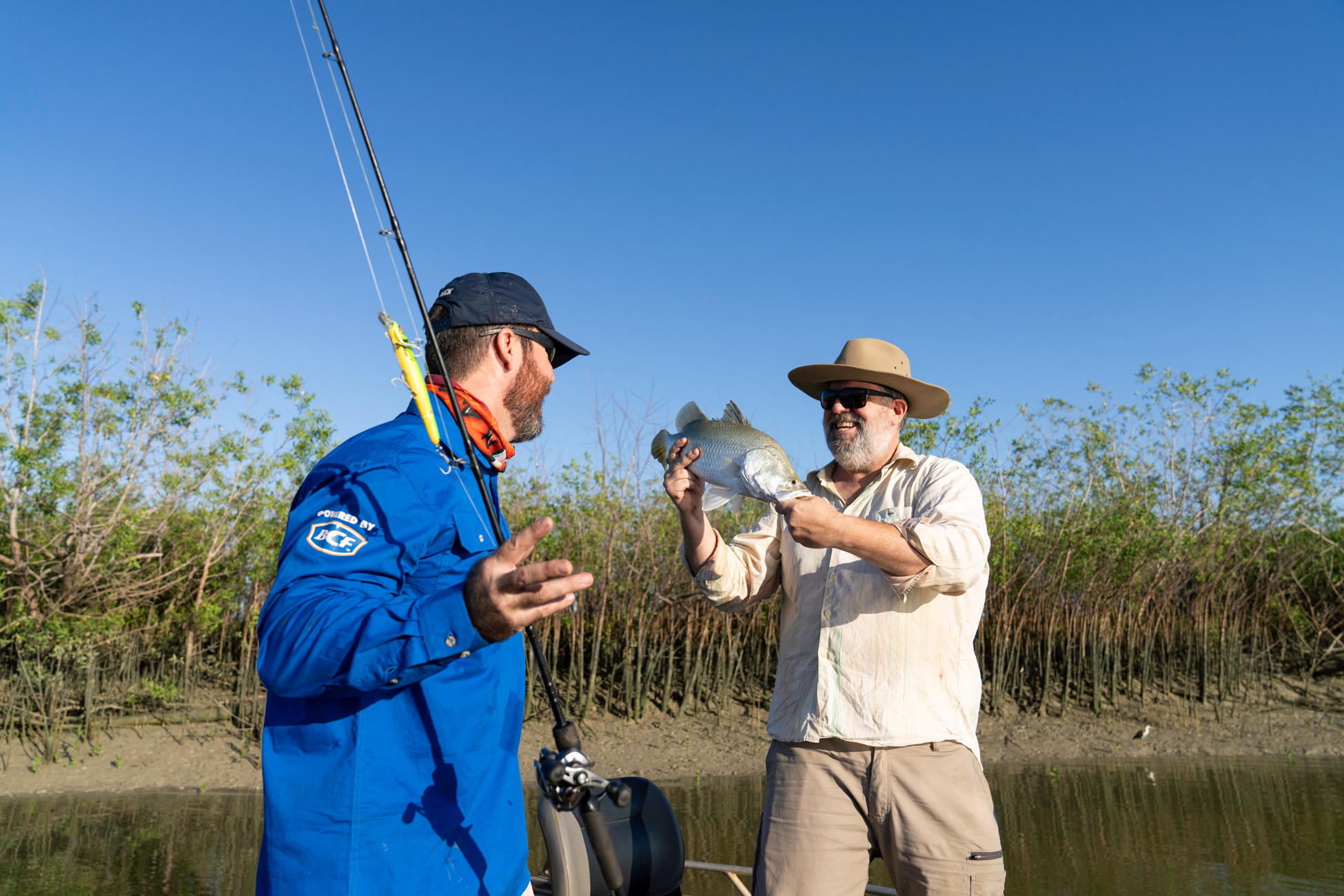 Two men on a boat, one in a blue shirt holding a fishing rod, the other in a hat holding a fish, with trees and a clear blue sky behind them.