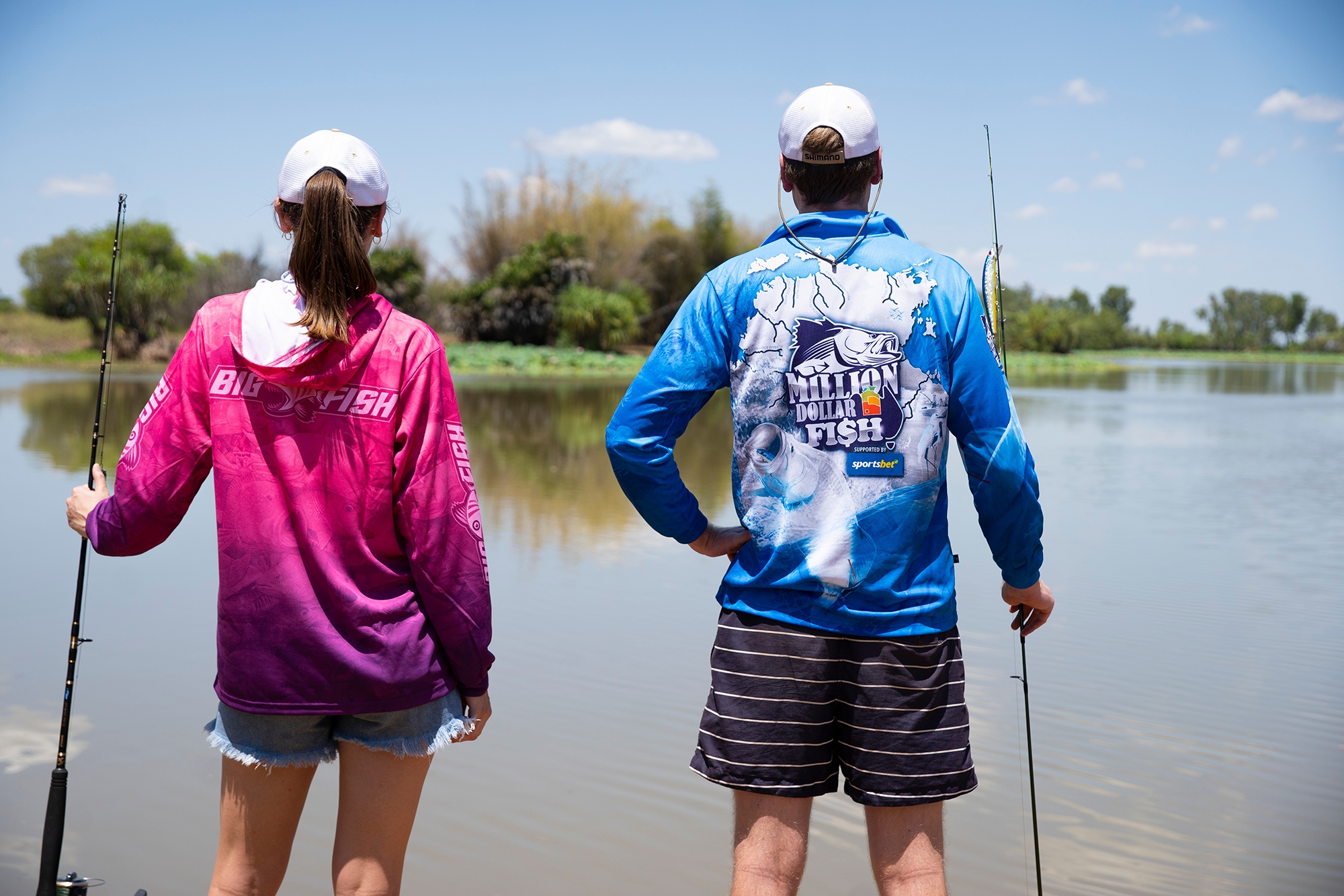 Two people in colorful fishing shirts stand by a calm lake, holding fishing rods, with trees and a clear sky in the background.