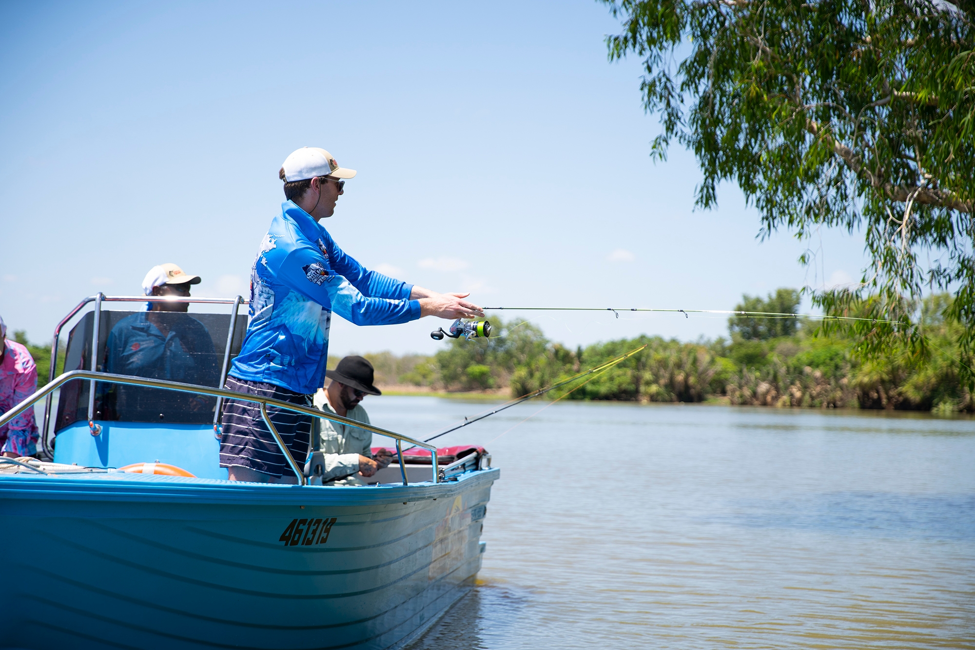 Three people fishing from a blue boat on a calm river, surrounded by lush greenery under a clear blue sky.