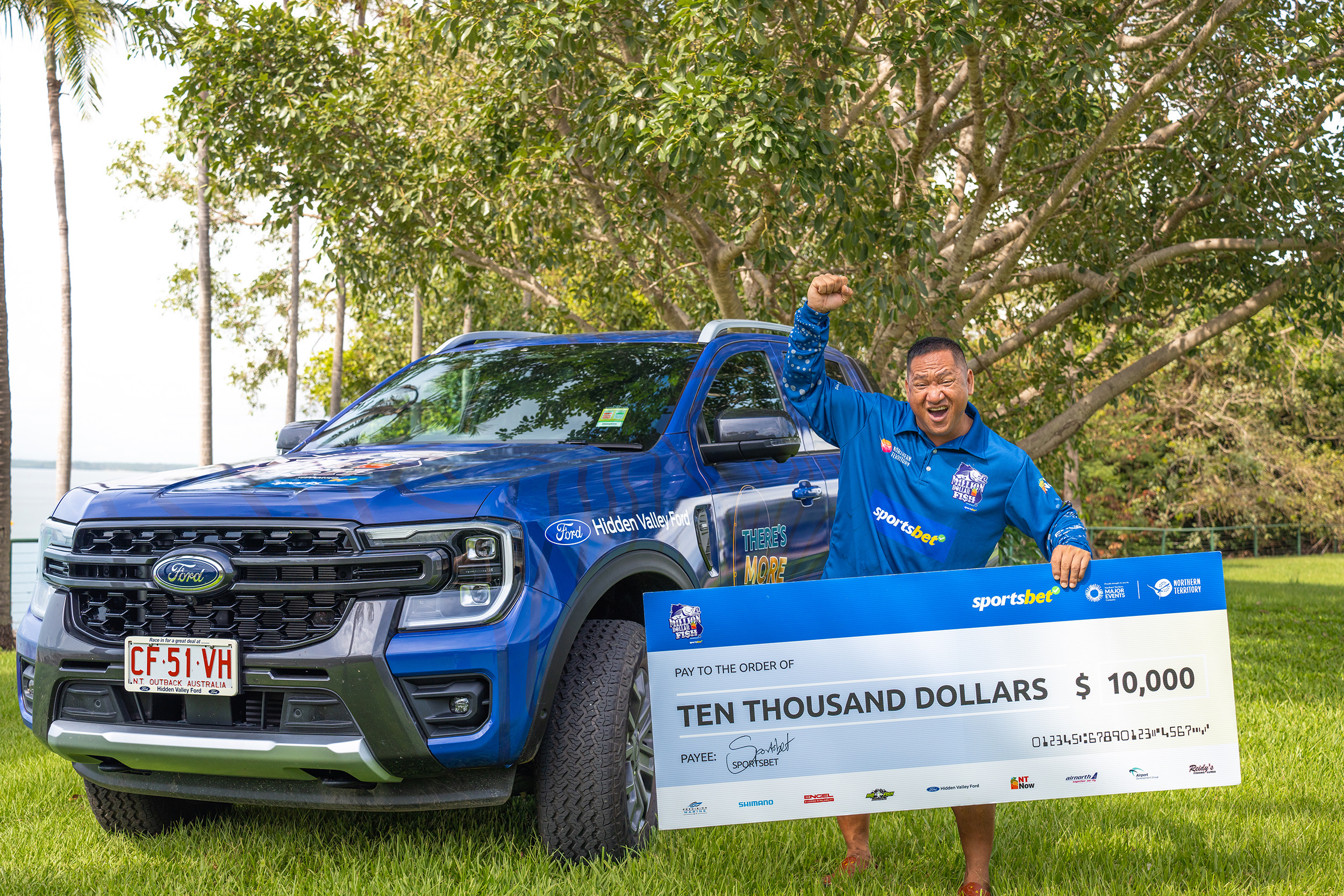 Man in a blue shirt celebrates holding a large $10,000 check in front of a blue Ford truck, with trees in the background.