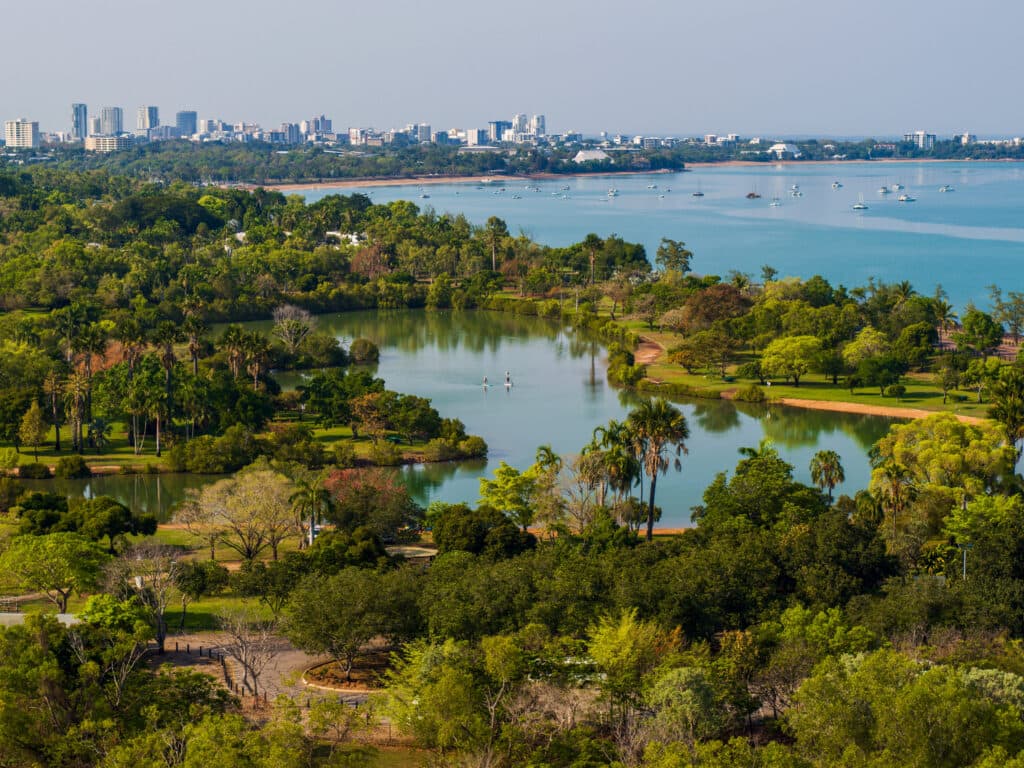 Aerial view of a lush, green park with a lake, surrounded by trees, near a coastal city skyline under a clear blue sky.
