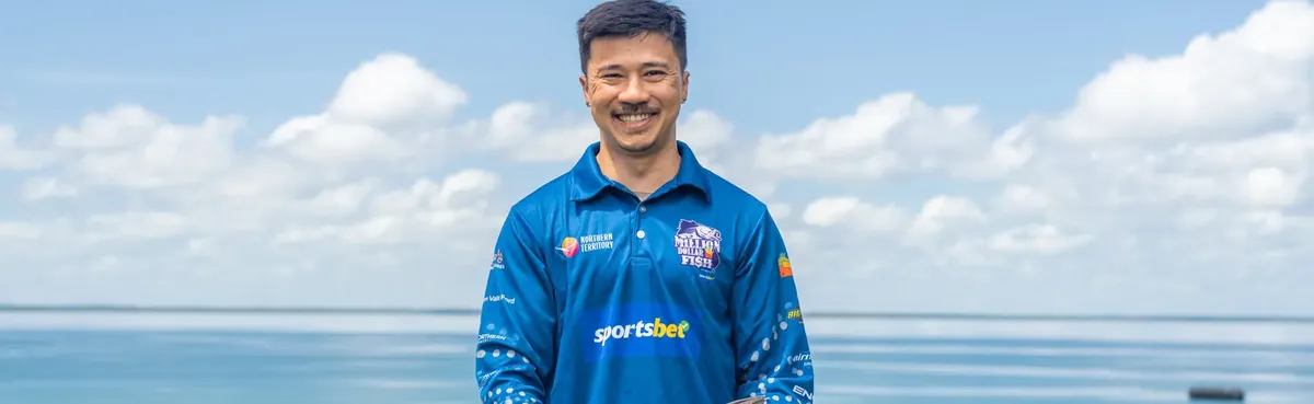 Smiling man in a blue sports jersey stands outdoors with a cloudy sky and water in the background.
