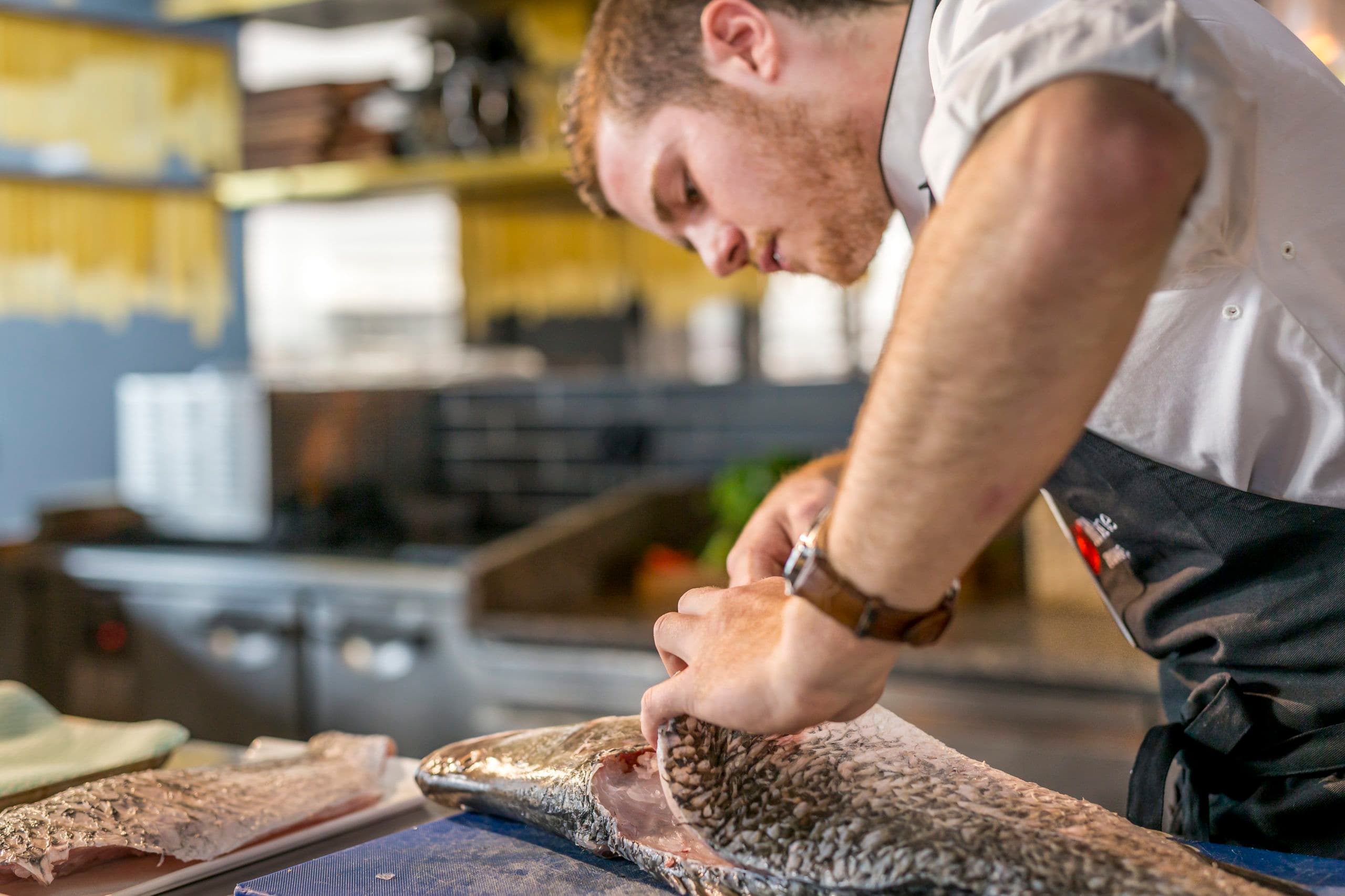 Chef filleting a large fish on a blue cutting board in a professional kitchen setting.