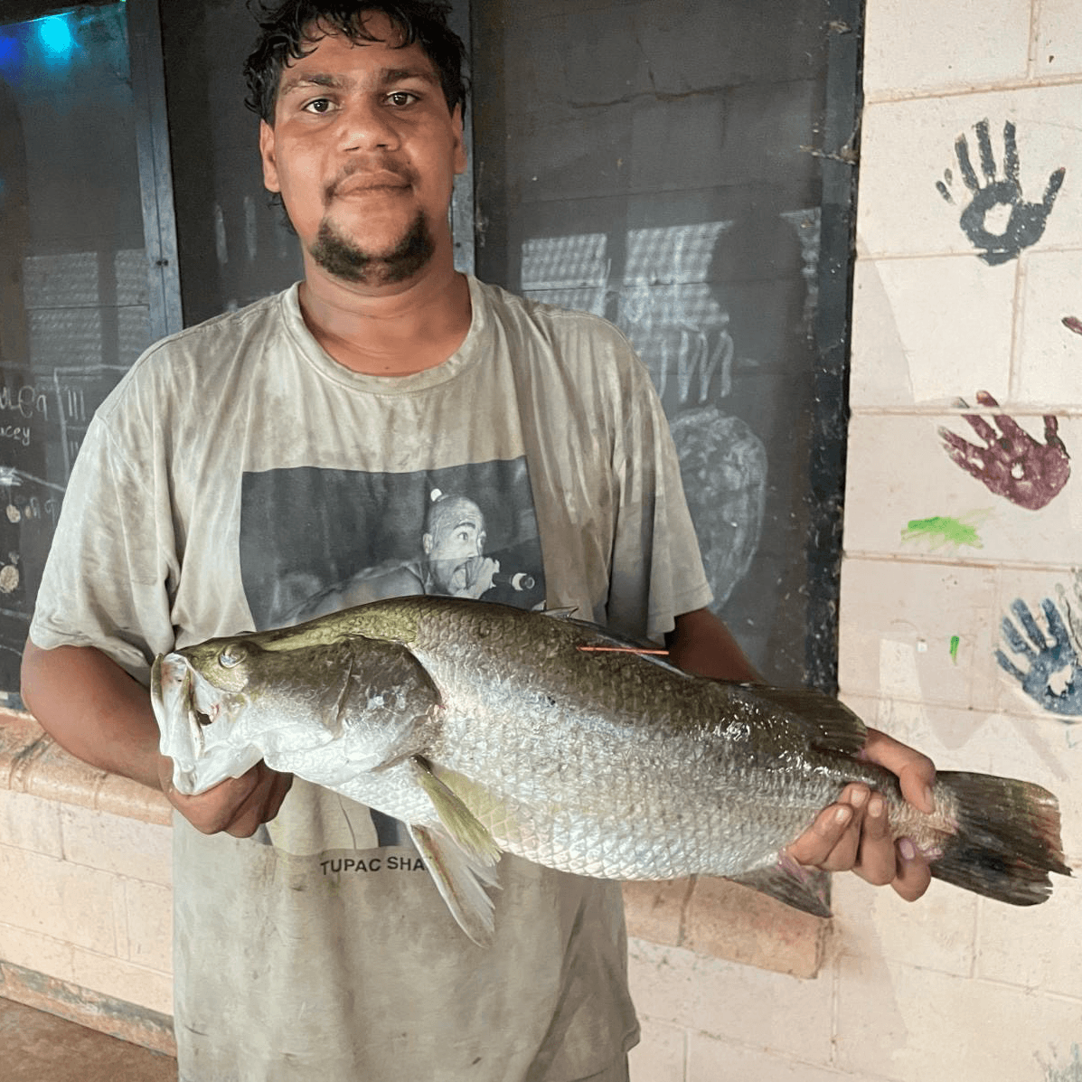 Man holding a large fish, standing in front of a wall with colorful handprints and a window. He is wearing a T-shirt with a graphic print.