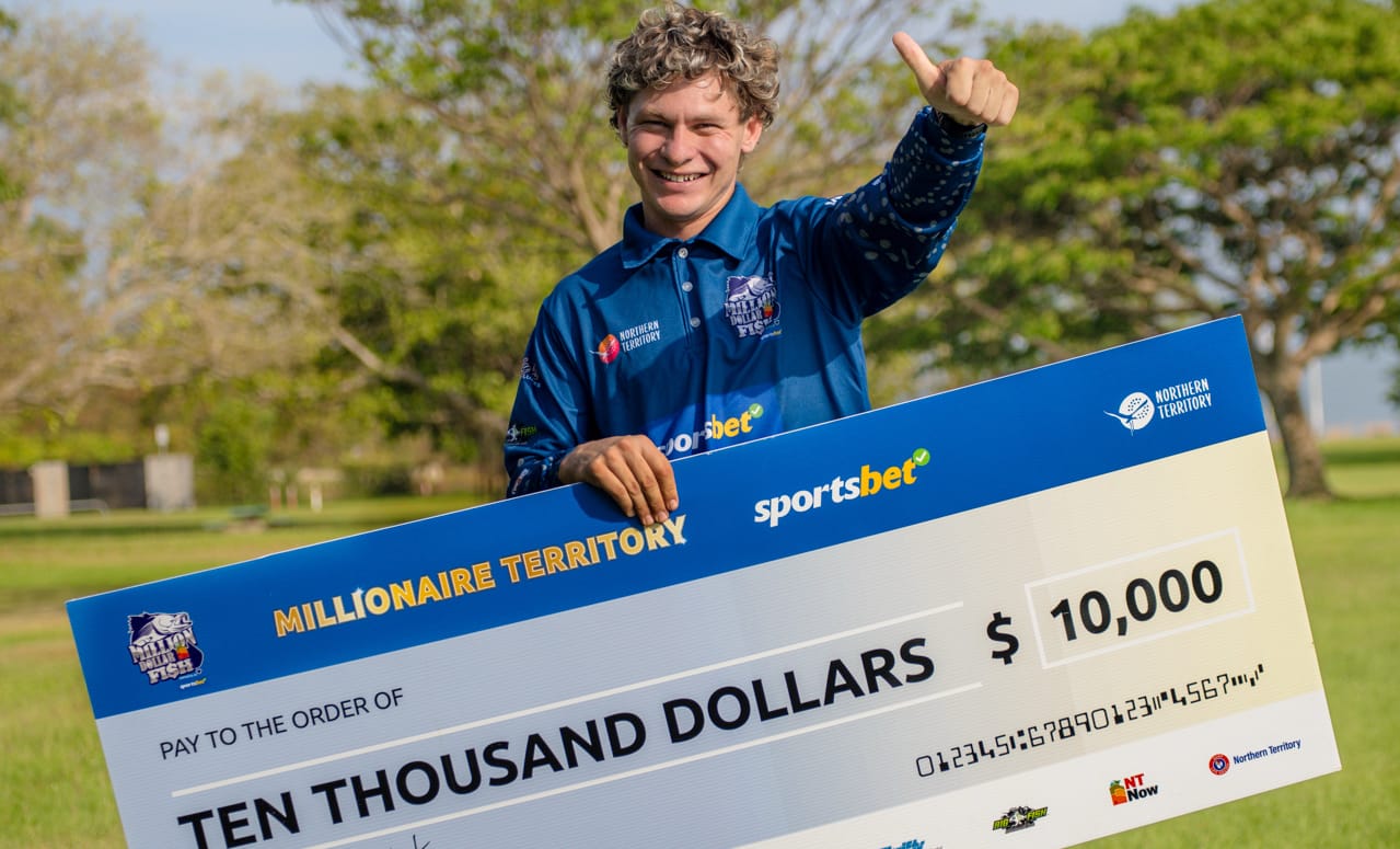 Person smiling and giving a thumbs-up while holding a large $10,000 check outdoors, surrounded by trees.
