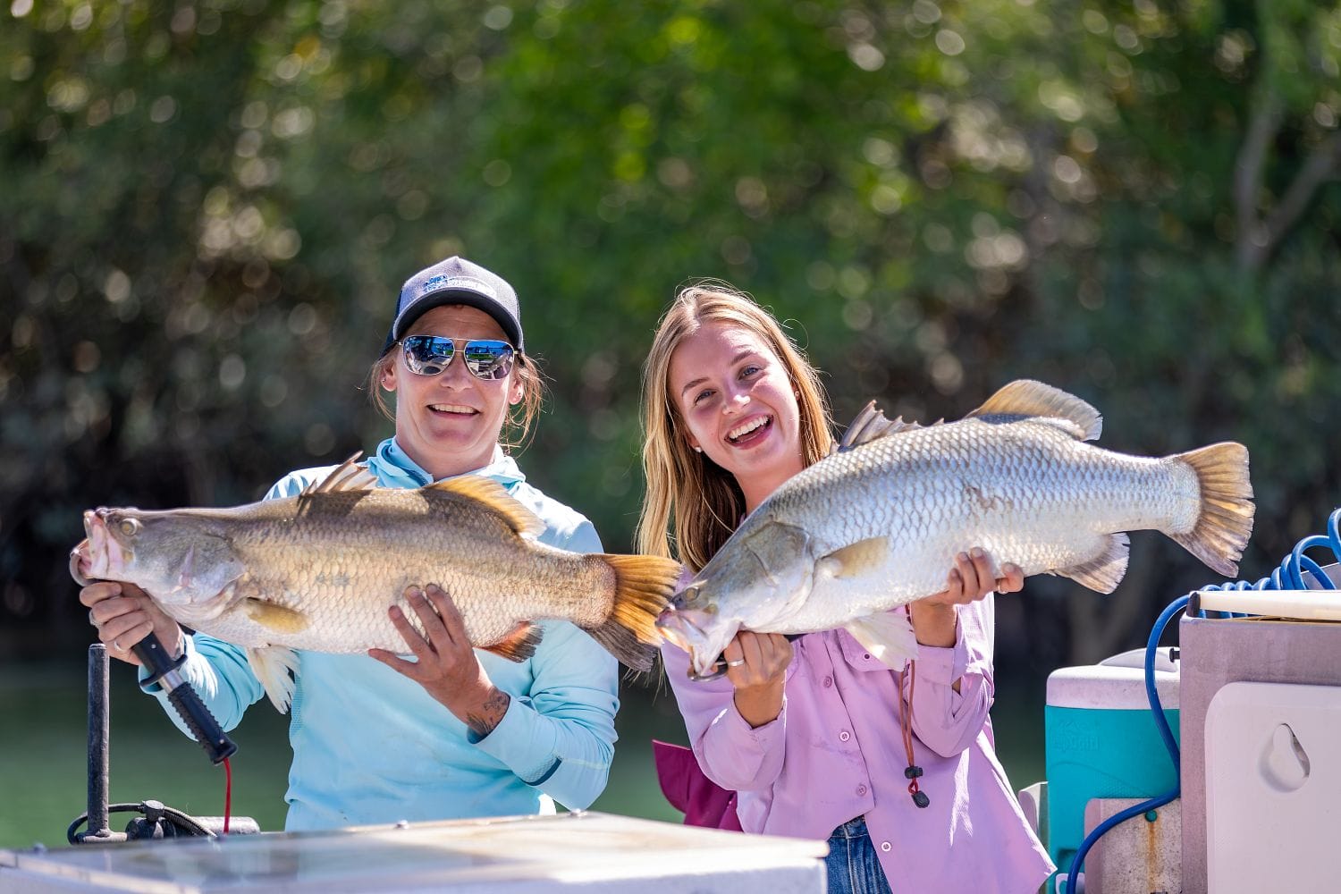 Two people smiling and holding large fish on a boat, with trees in the background. Both appear to be enjoying a successful fishing trip.