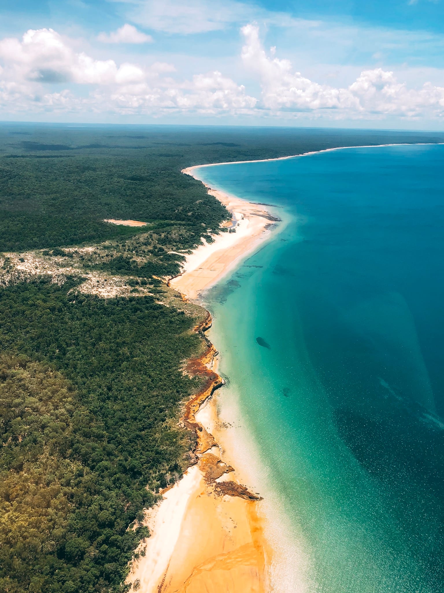 Aerial view of a coastline with turquoise waters, golden sandy beaches, and lush green forest under a bright blue sky with scattered clouds.