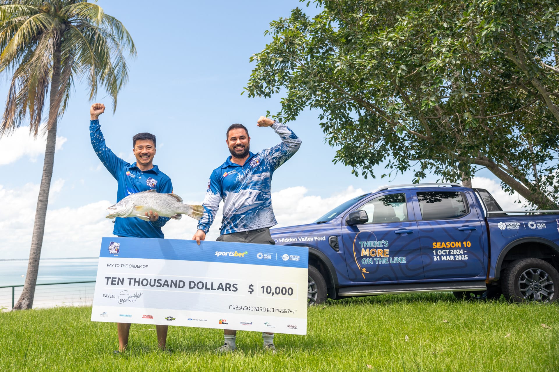 Two men celebrate with a large fish and a $10,000 check. A blue truck and palm tree are in the background under a clear sky.