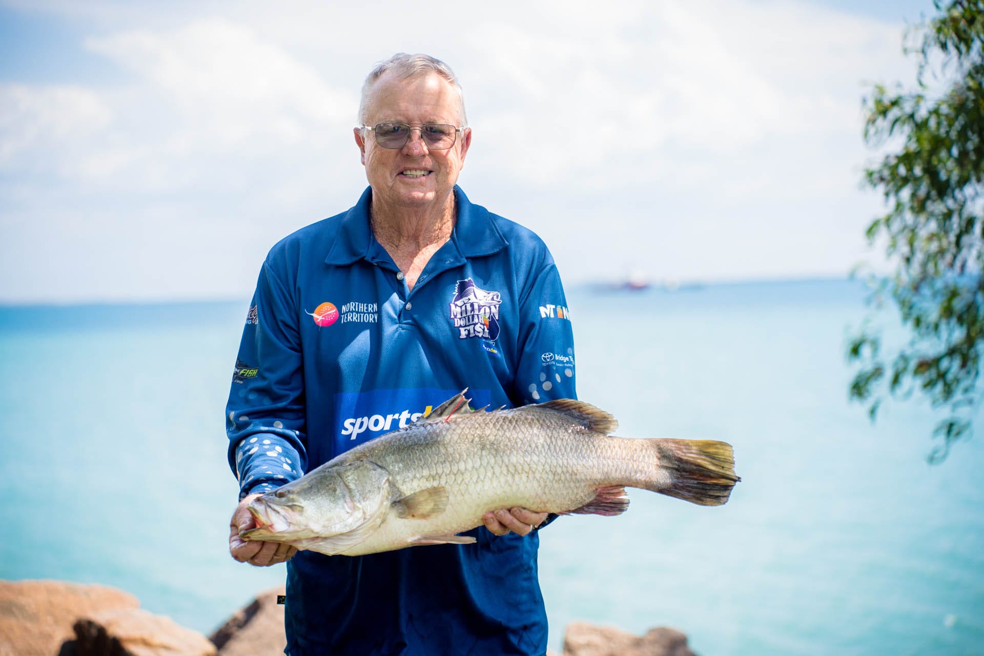 Man holding a large fish by a lake, wearing a blue sports shirt. Bright, sunny day with clear blue water and sky in the background.