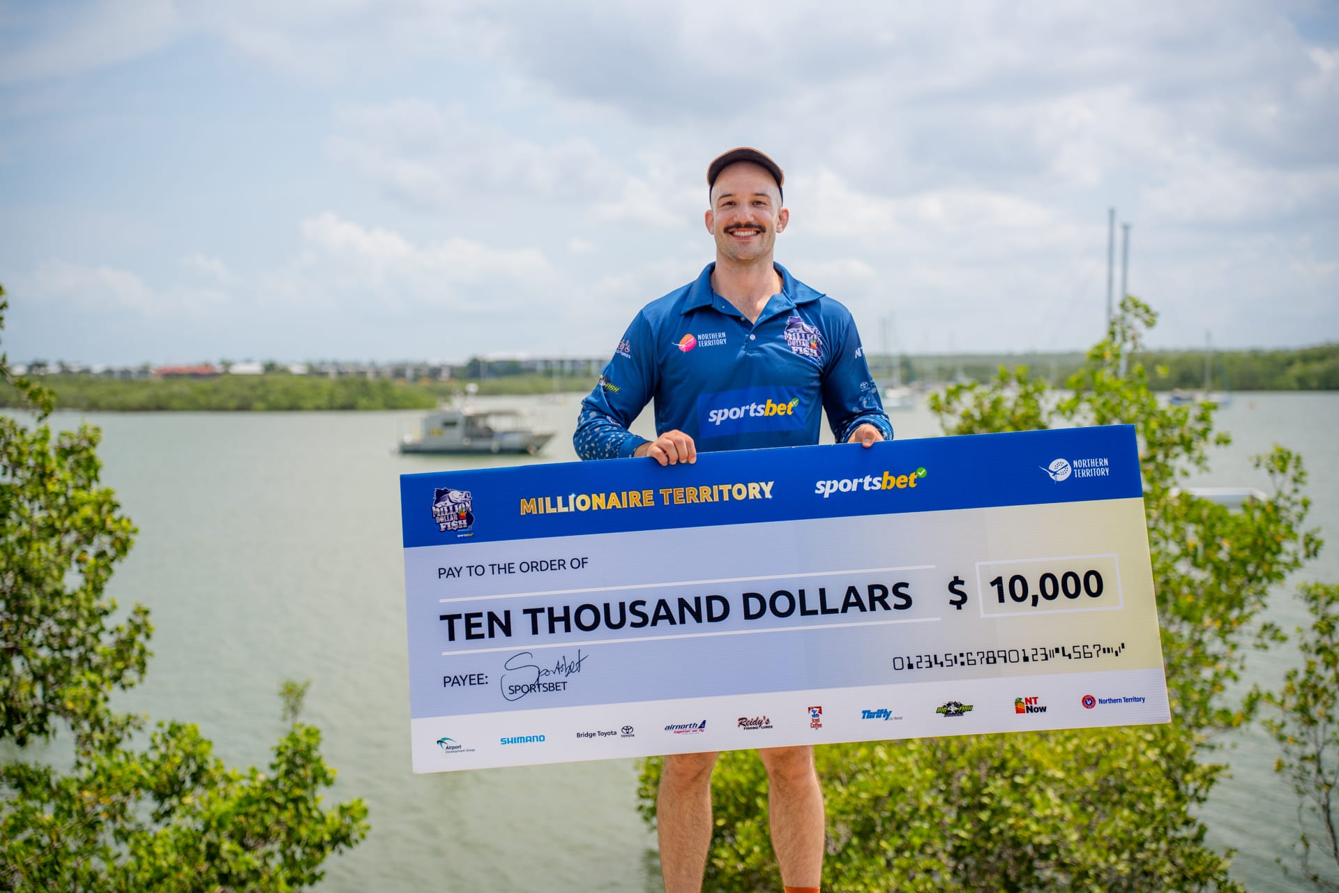 A person holding a large $10,000 check from Sportsbet, standing outdoors with water and trees in the background.