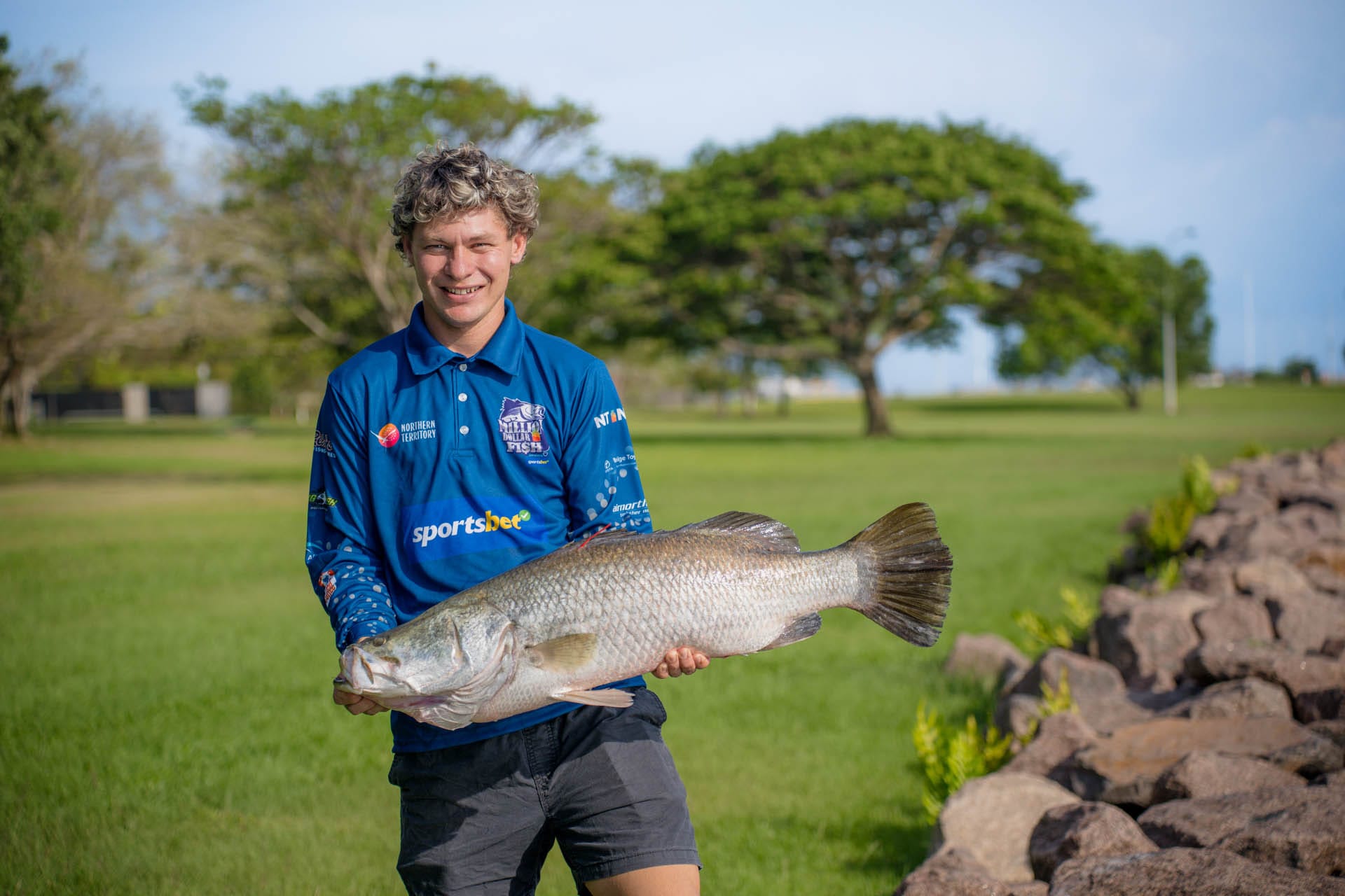 Person holding a large fish outdoors, wearing a blue shirt, with green grass and trees in the background.