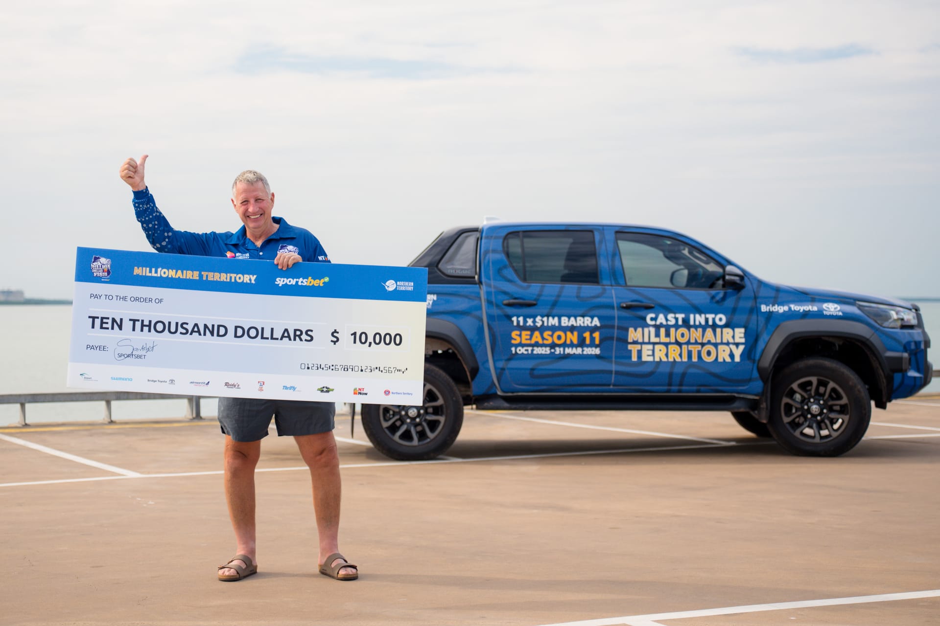 Man holding a large check for $10,000, standing on a pier with a branded truck in the background.