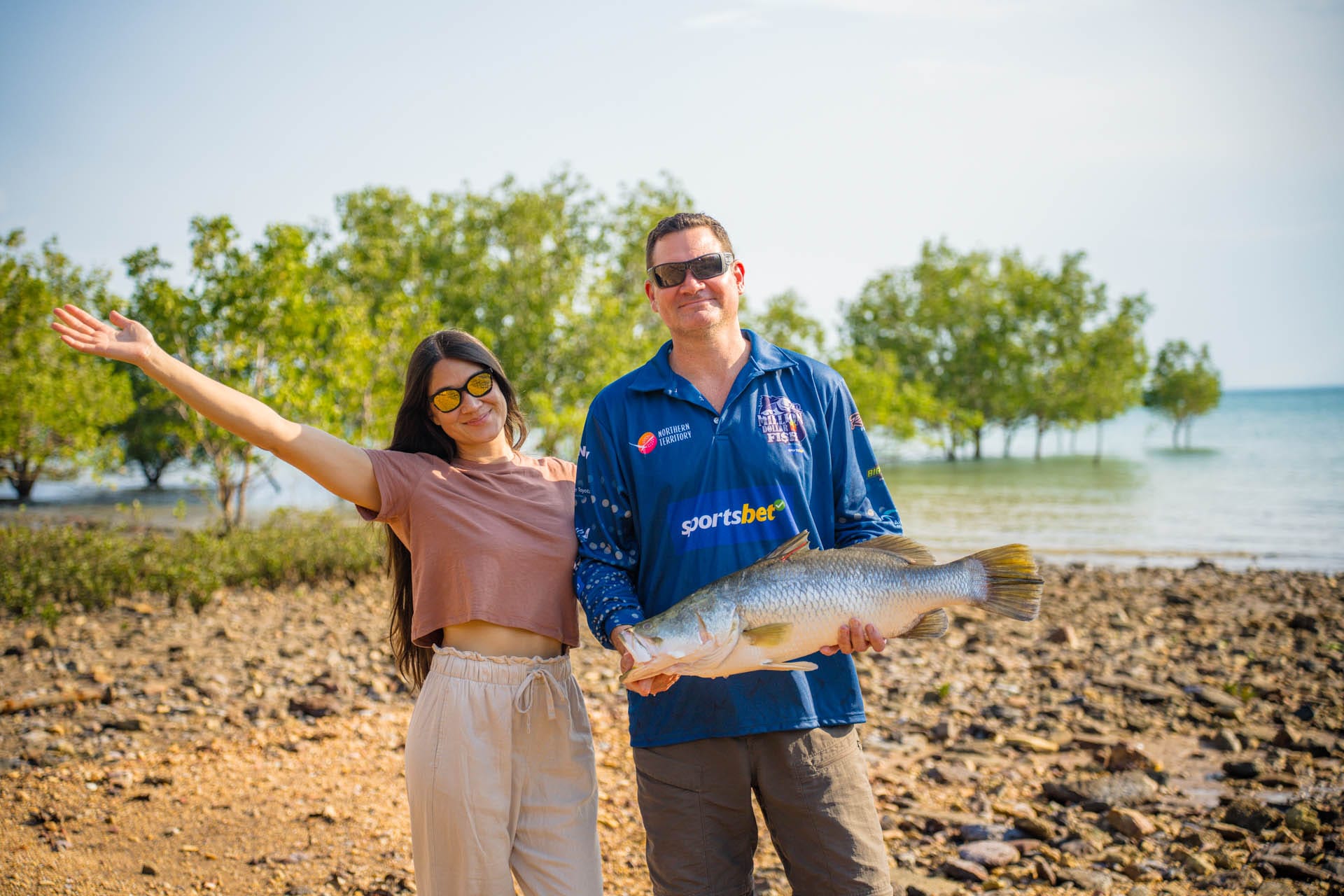 A smiling couple stands on a rocky shore. The man holds a large fish. Trees and water are visible in the background.