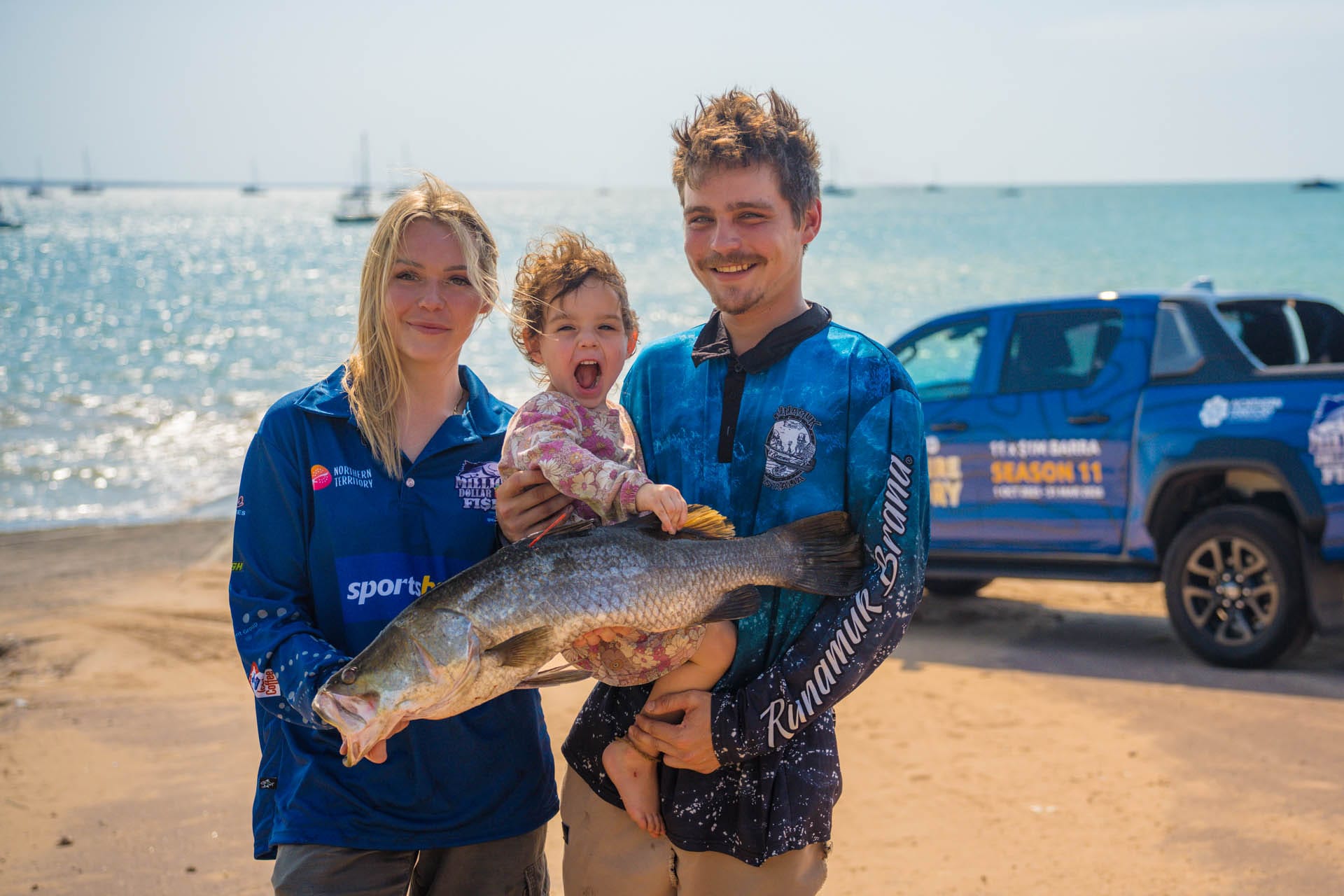 A family on a sunny beach holds a large fish. The woman and man wear blue shirts, while the child sits atop the fish, smiling widely.