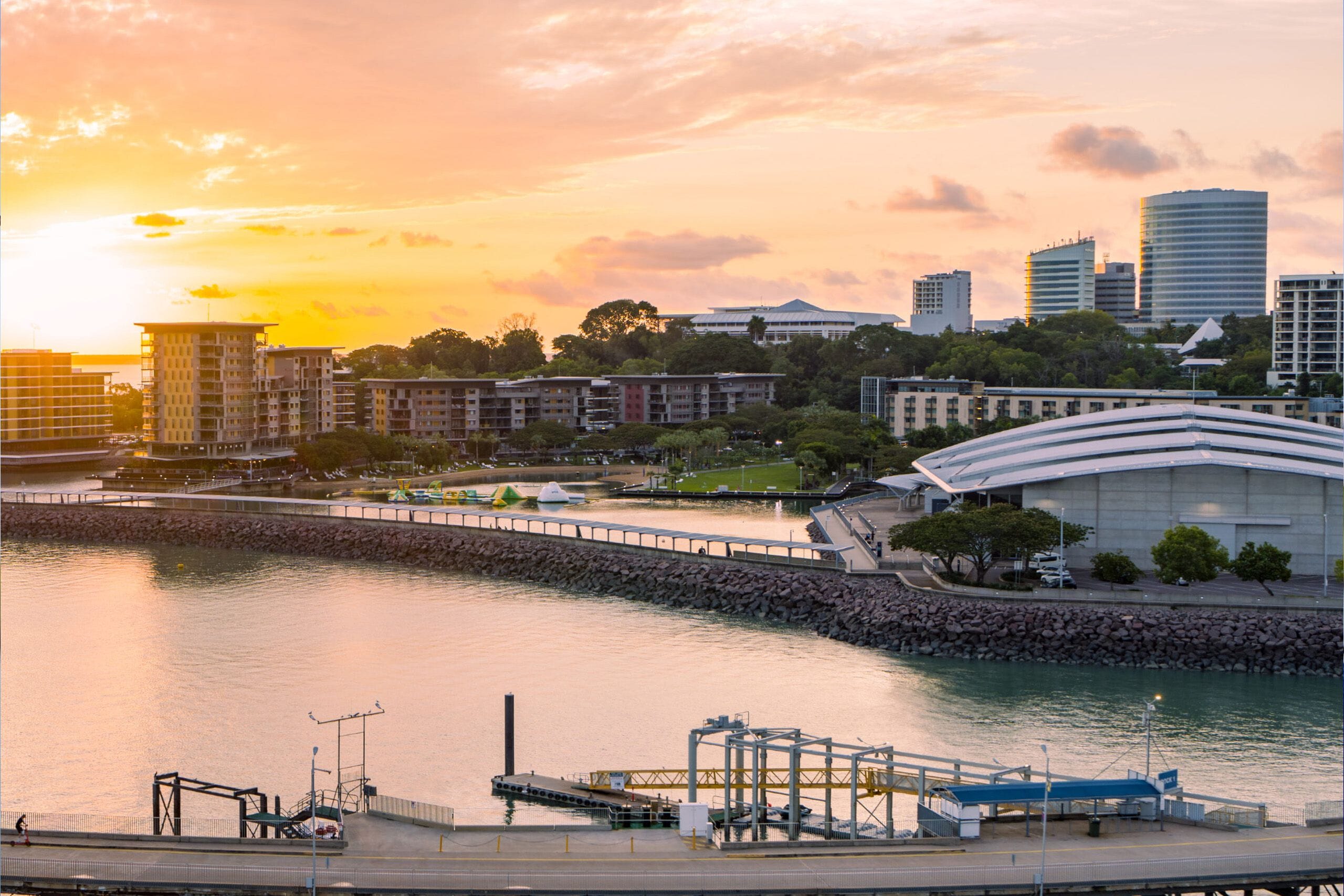 Sunset view of a waterfront cityscape with modern buildings, a park, and calm water reflecting the colorful sky.