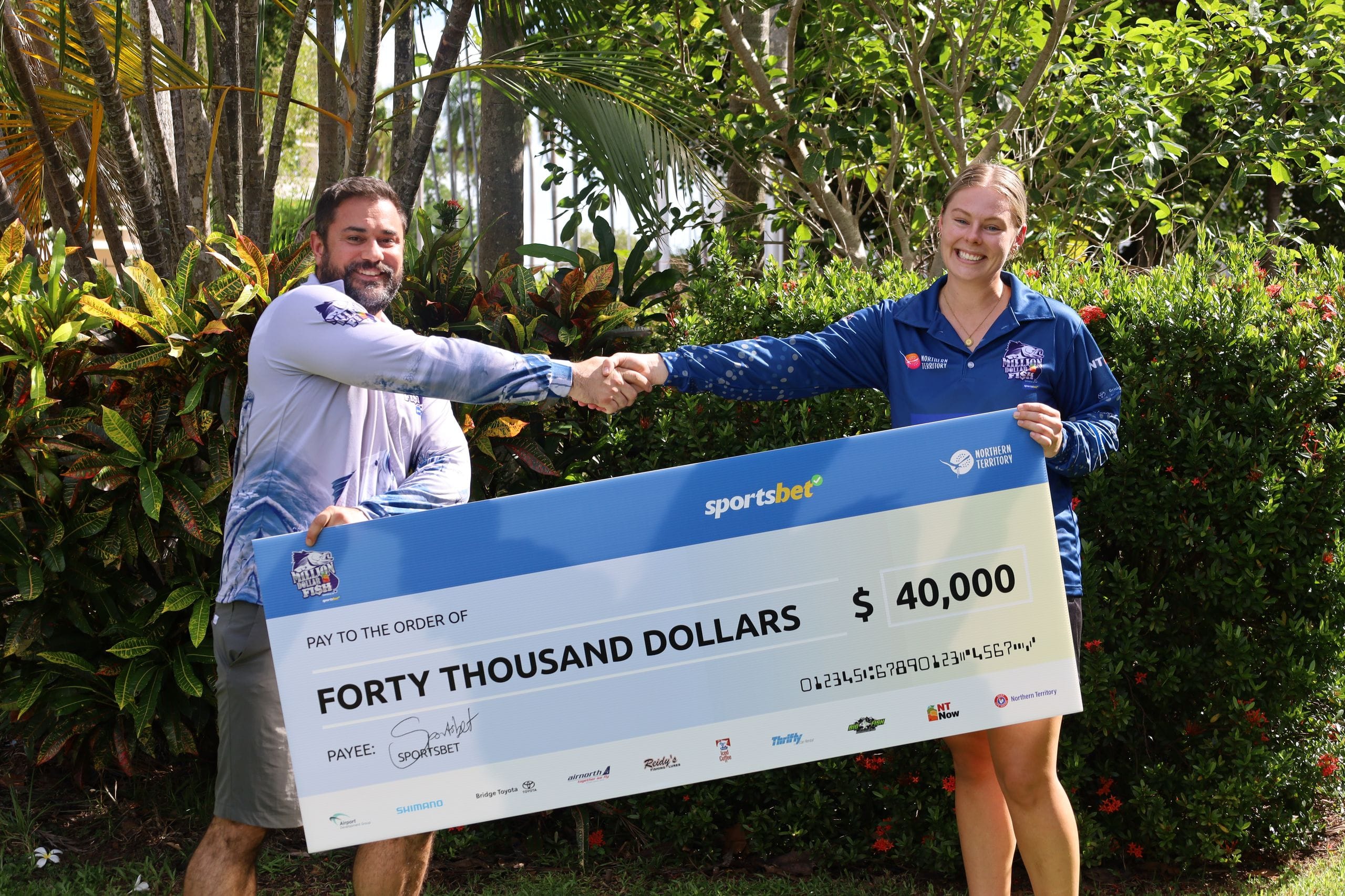 Two people shaking hands while holding a large check for forty thousand dollars in a garden setting.
