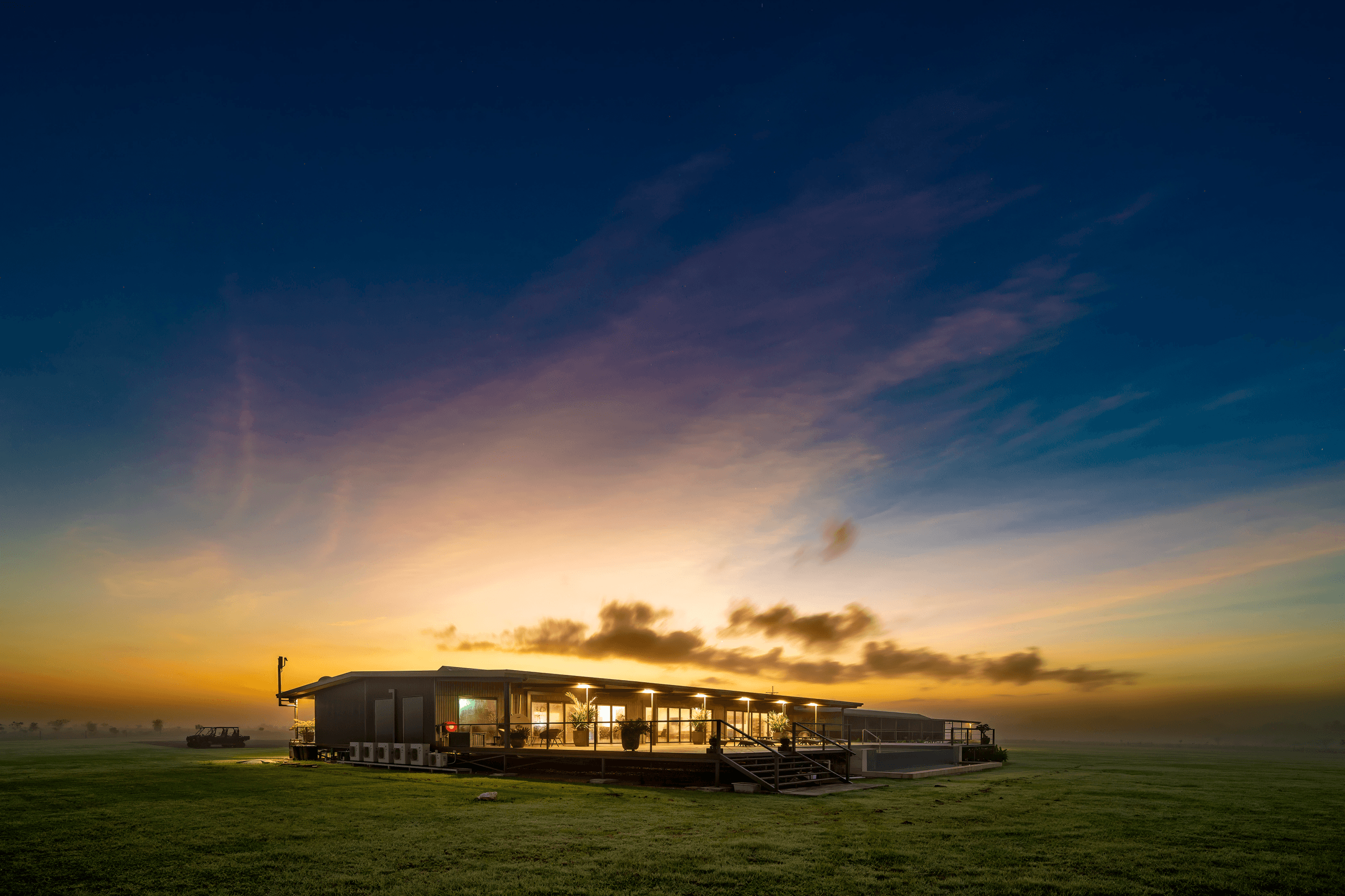 A modern building with large windows is illuminated against a vibrant sunset sky, surrounded by an expansive grassy field.