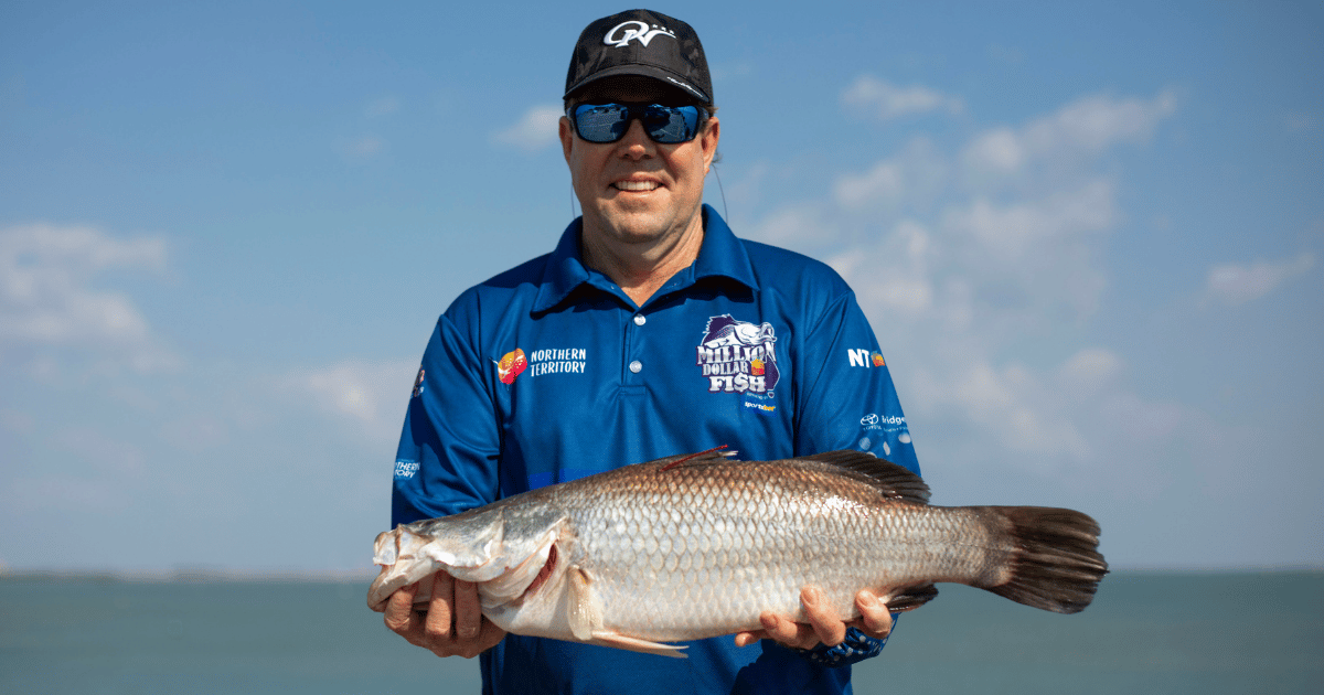 A man in a blue Million Dollar Fish shirt and cap holds a large barramundi, with the ocean and blue sky in the background.