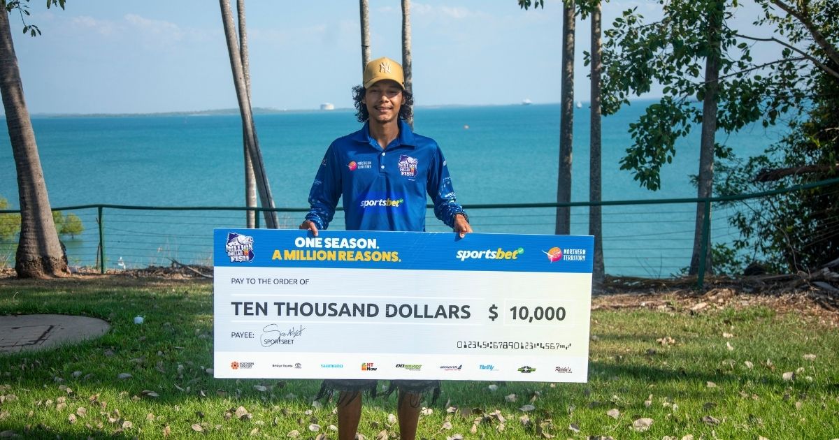 A man in a blue Million Dollar Fish shirt holds an oversized $10,000 cheque outdoors, with tropical trees and the ocean in the background.