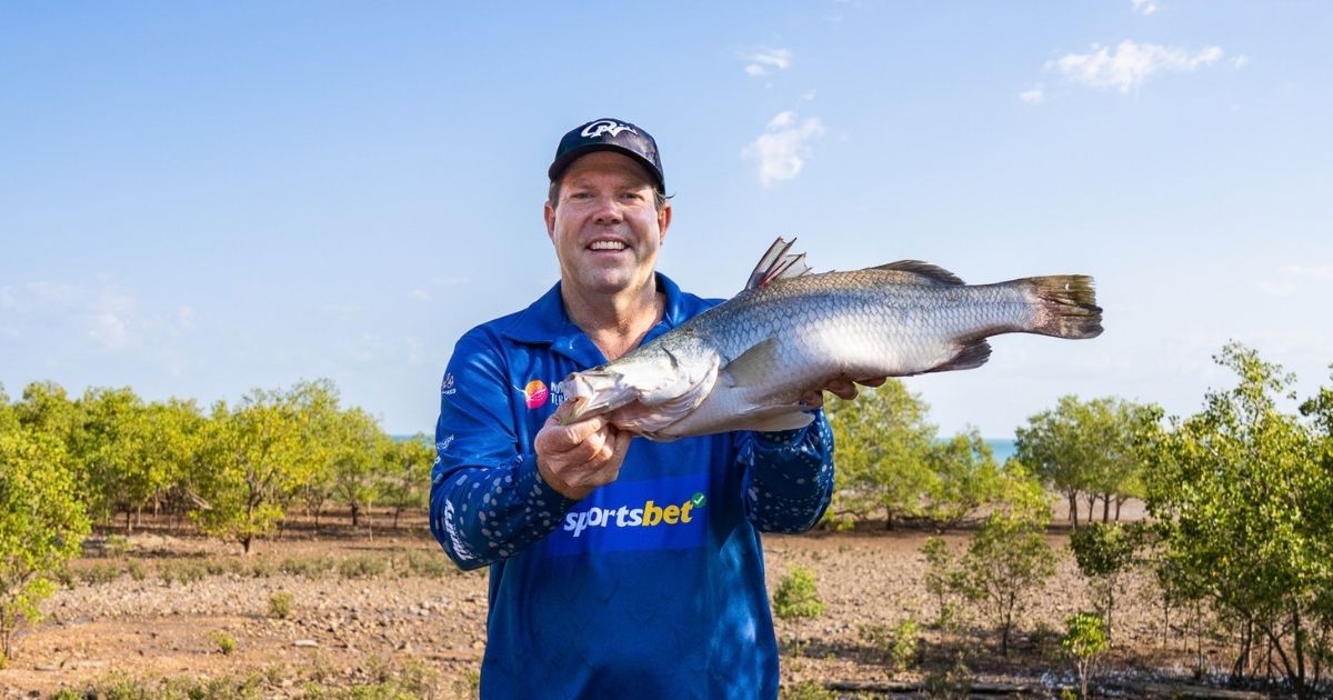 A man wearing a blue fishing shirt and cap holds a large barramundi outdoors, with mangroves and a clear blue sky in the background.