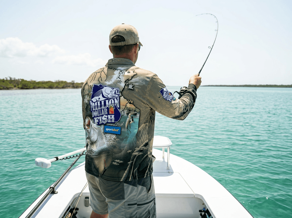 A person standing on the bow of a small fishing boat casts a fishing line across calm turquoise water. The person is wearing a long-sleeve Million Dollar Fish fishing shirt, viewed from the back, featuring a large Million Dollar Fish logo, SportsBet branding, a map of the Northern Territory, and artwork depicting a barramundi, wetlands, birds and waterways. The boat is surrounded by open water, with a low mangrove-lined shoreline visible in the distance under a bright sky. The image showcases the Million Dollar Fish apparel in an on-water recreational fishing setting.