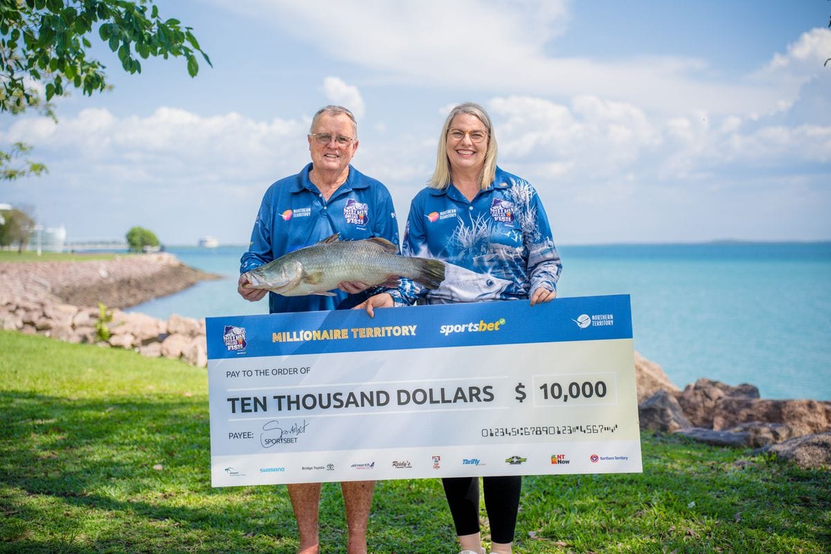 A man and woman smiling, holding a large fish and a $10,000 check, standing on grass with the ocean and clouds in the background.