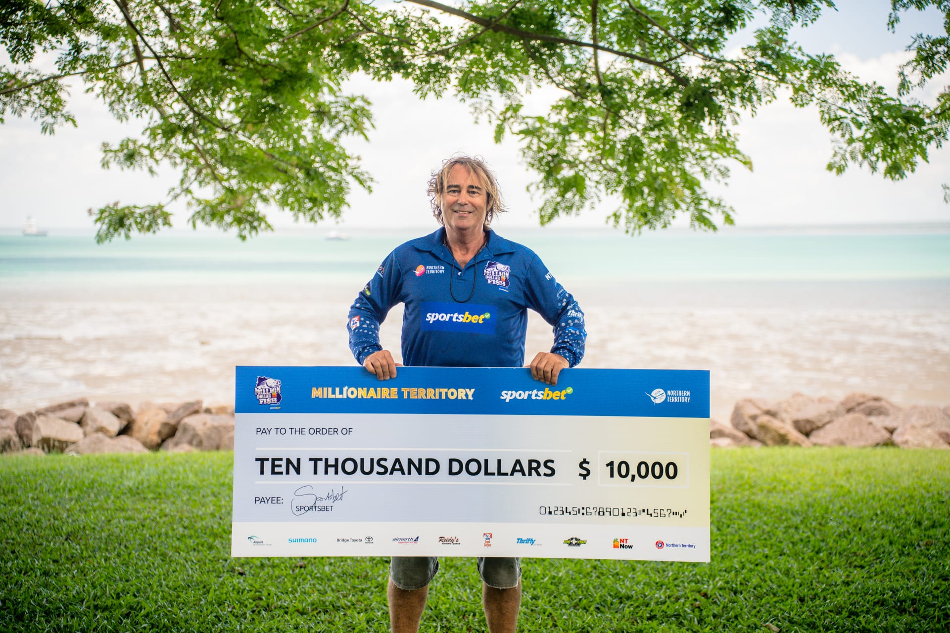 Person holding a large $10,000 check outdoors, standing on grass with a beach and ocean in the background.