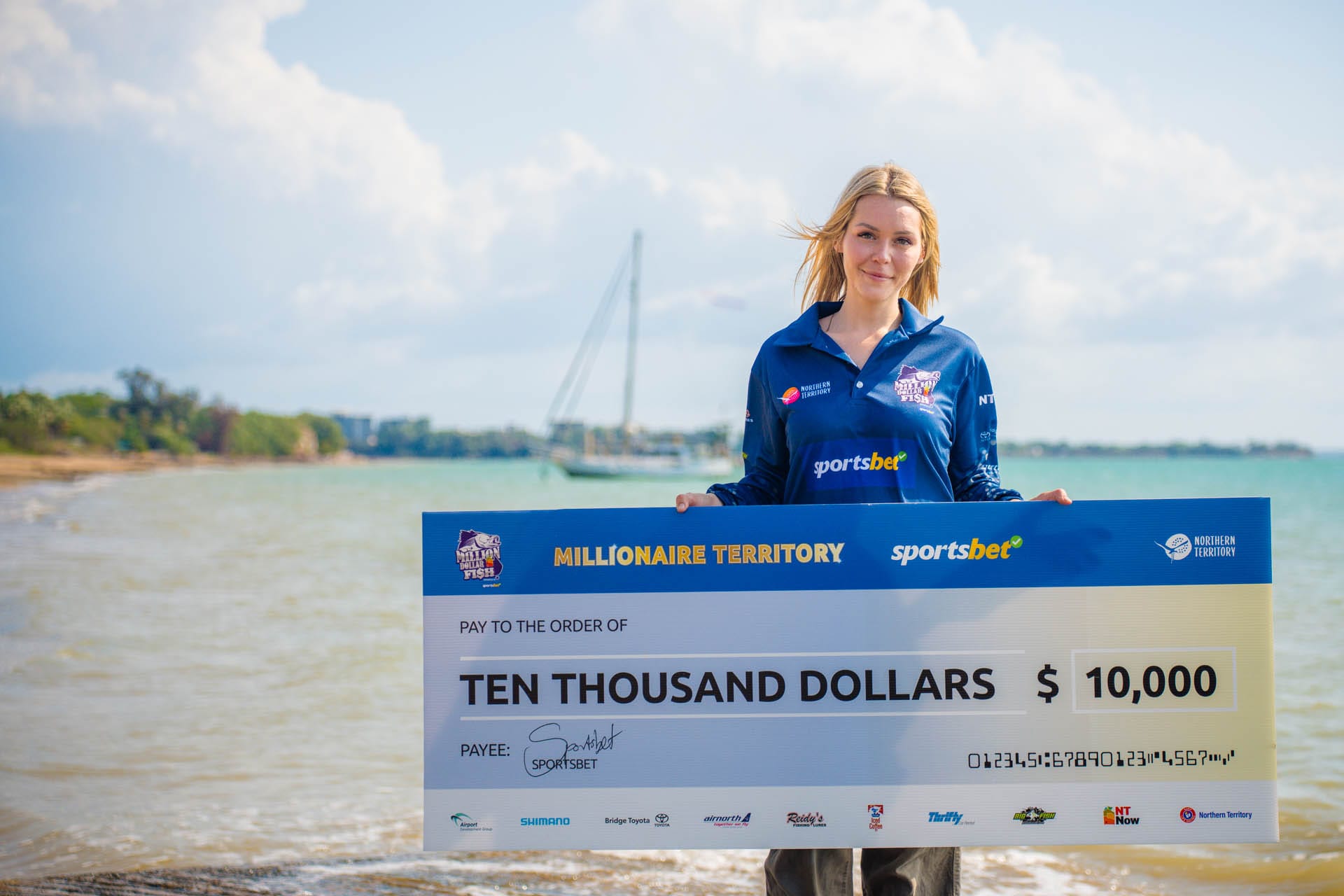 Woman holding a large $10,000 check by the sea, with a sailboat and cloudy sky in the background.