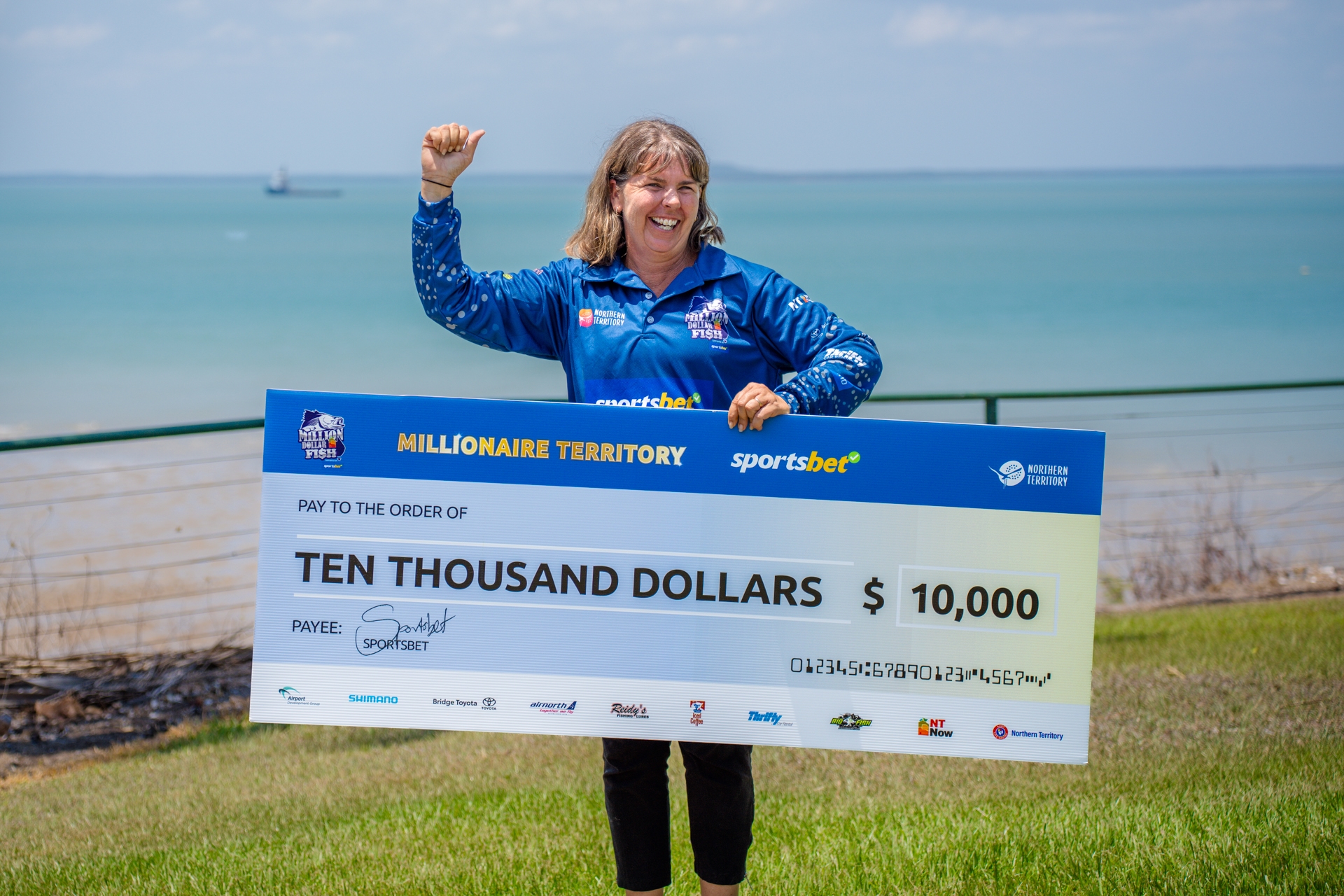 A woman in a blue shirt holds a large $10,000 check, smiling by the ocean with a ship in the background.