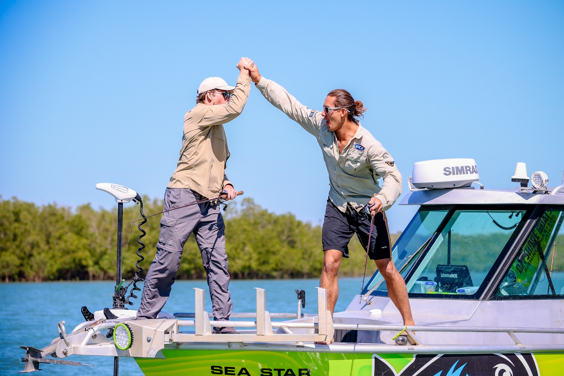 Two people on a boat high-five each other, both wearing casual outdoor clothing. The boat is on a calm body of water with trees in the background.