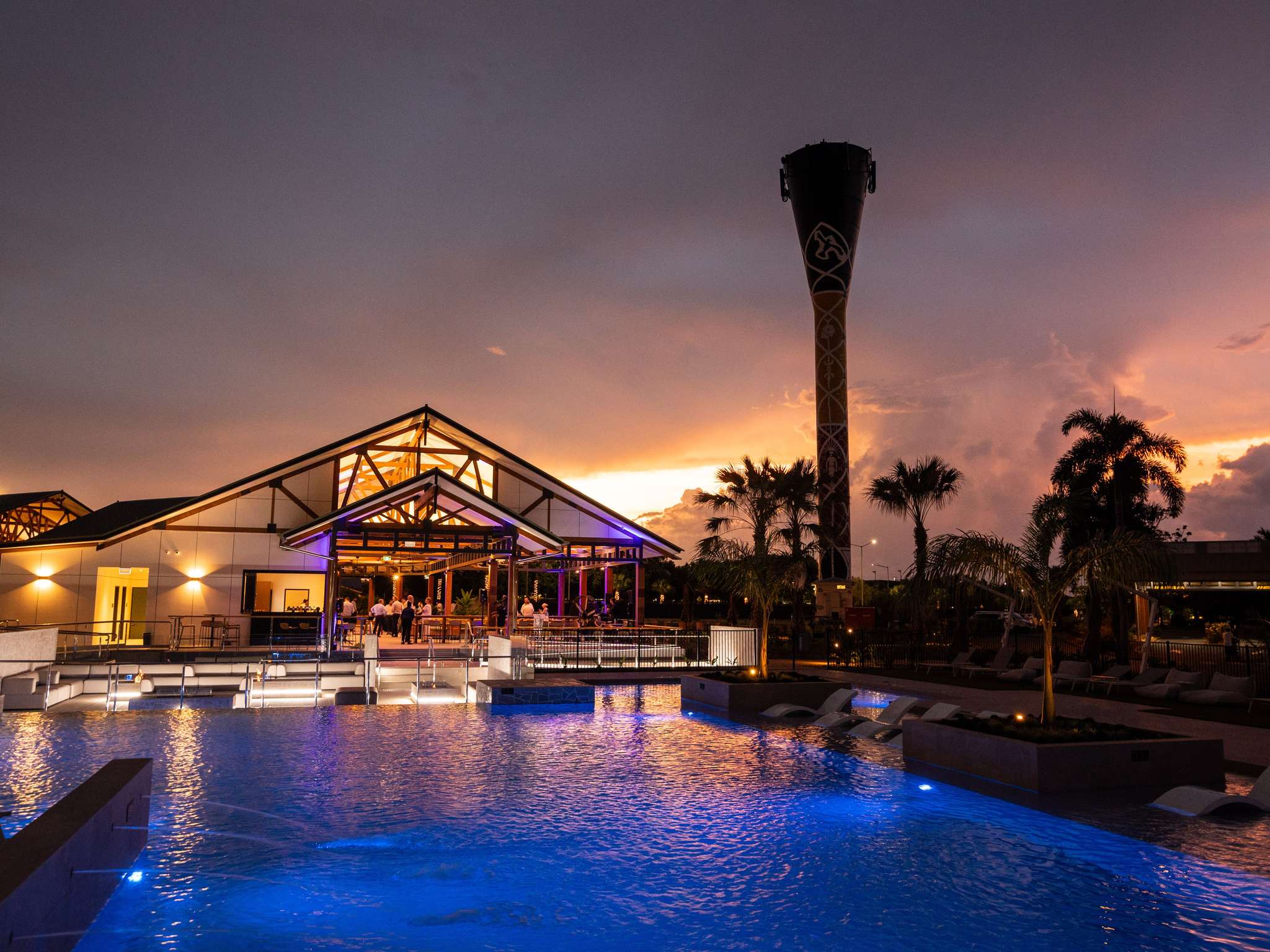 A modern building with large windows is illuminated at dusk beside a pool with blue lights. A tall tower and palm trees are visible in the background.