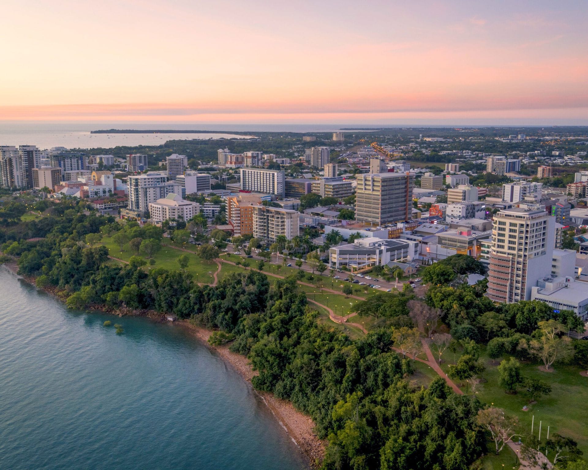 Aerial view of a coastal city at sunset, featuring high-rise buildings, green parks, and a calm shoreline under a pink-orange sky.