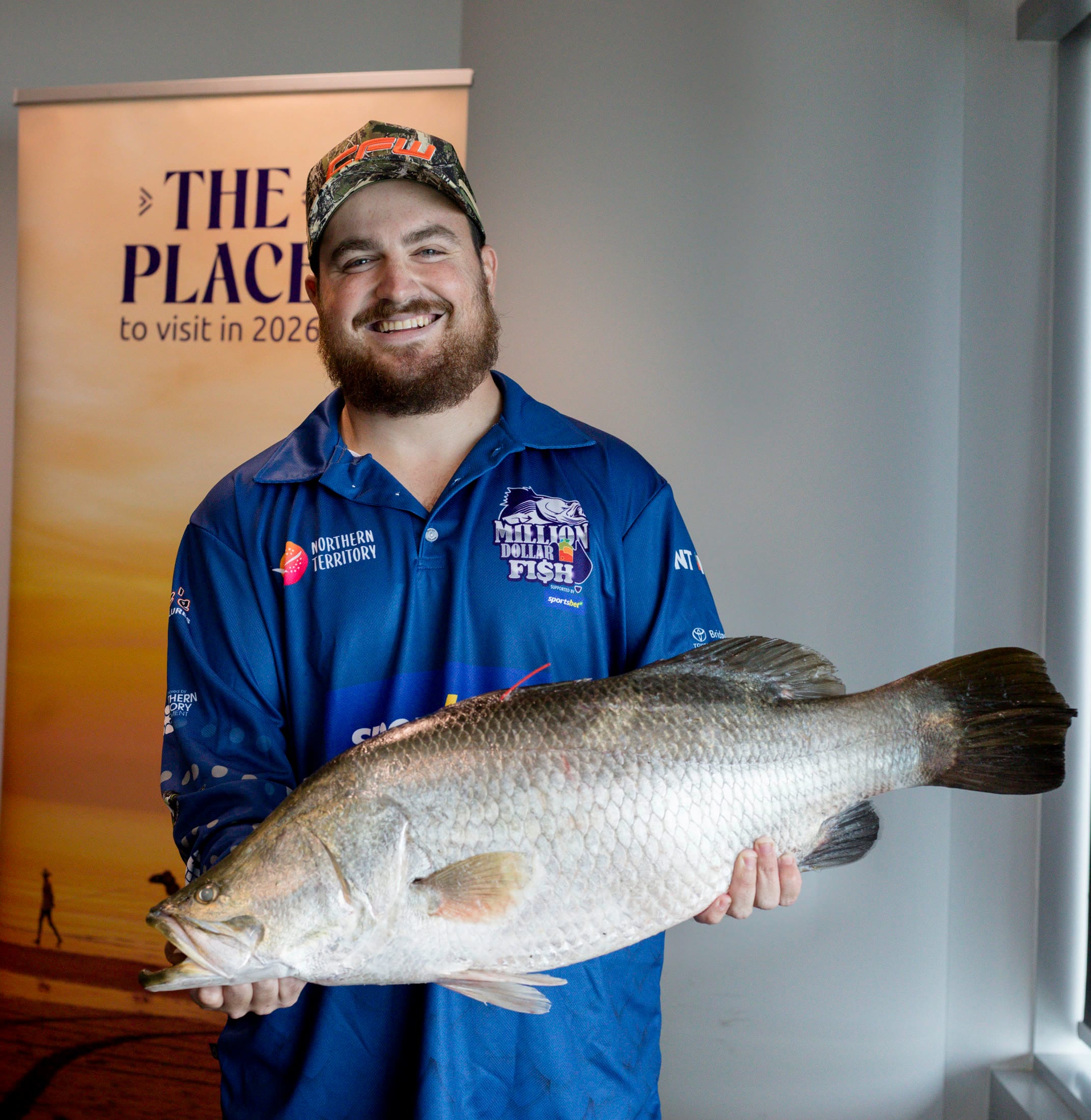 A man wearing a hat and a blue short that has a Million Dollar Fish logo is holding a large Barramundi fish with a red tag attached.