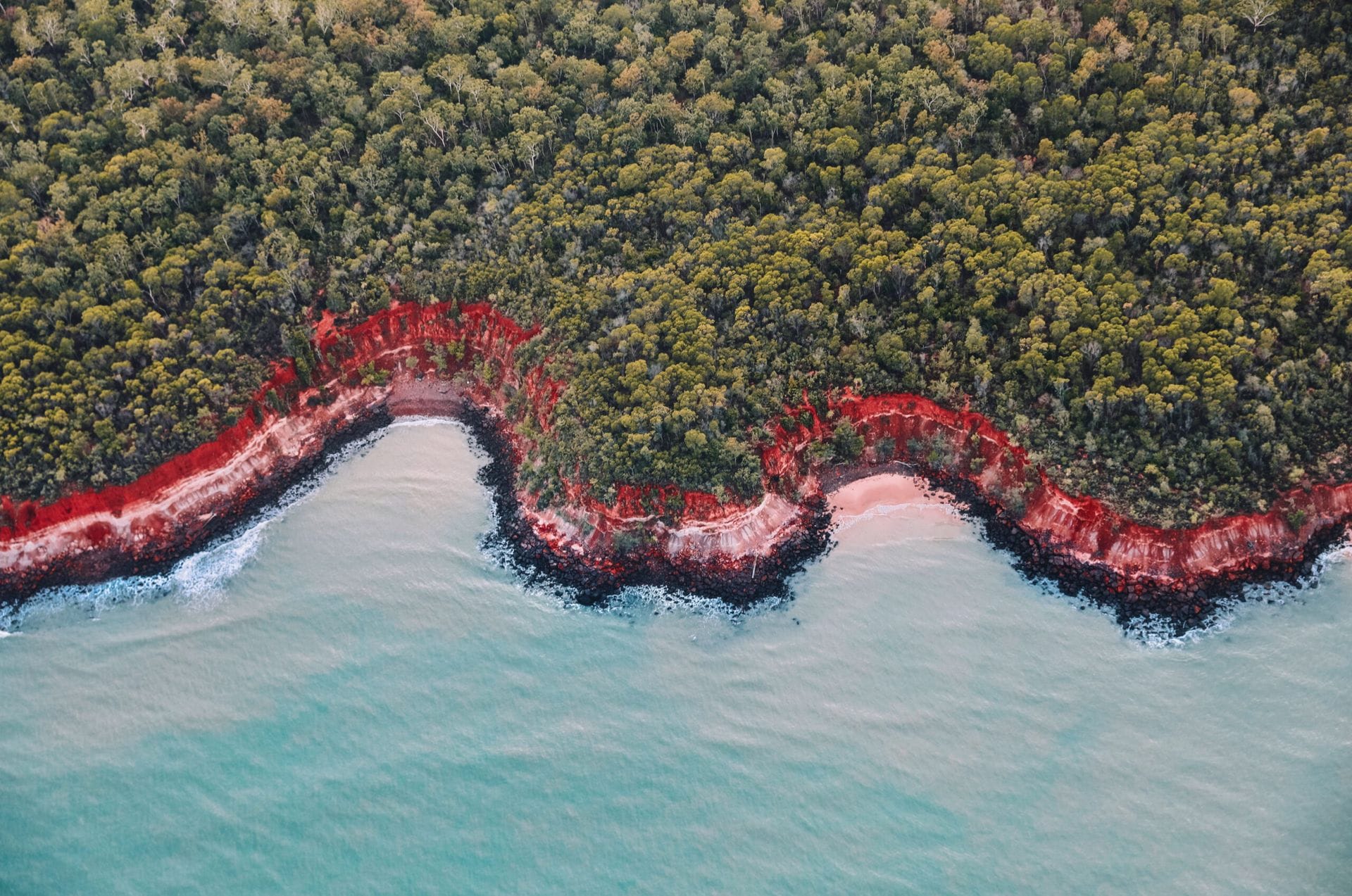 Aerial view of a coastal landscape with lush green forest, vibrant red cliffs, and turquoise ocean waves gently hitting the shore.