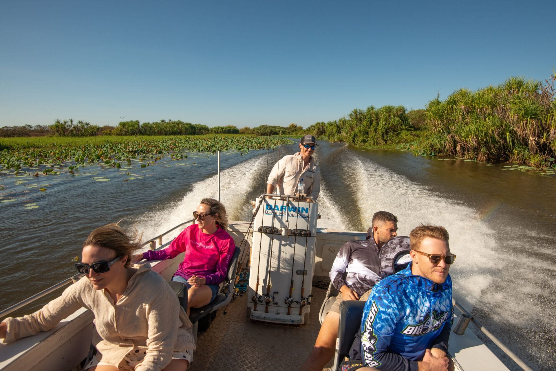 A group enjoys a high-speed boat ride through a lush, green wetland under a clear blue sky, with water splashing behind them.