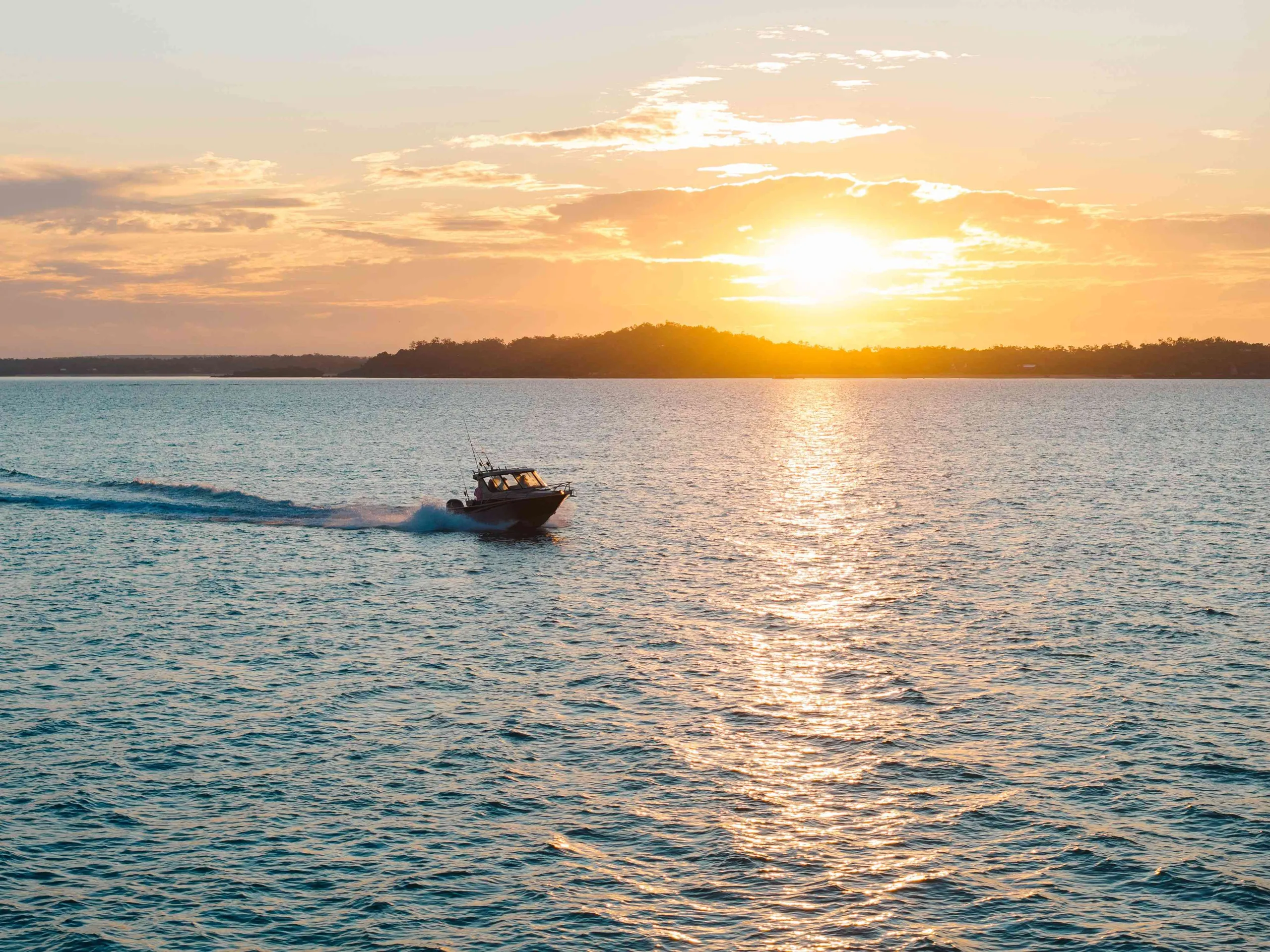A fishing boat cruises across the water at sunset, framed by golden reflections and a vibrant coastal sky.