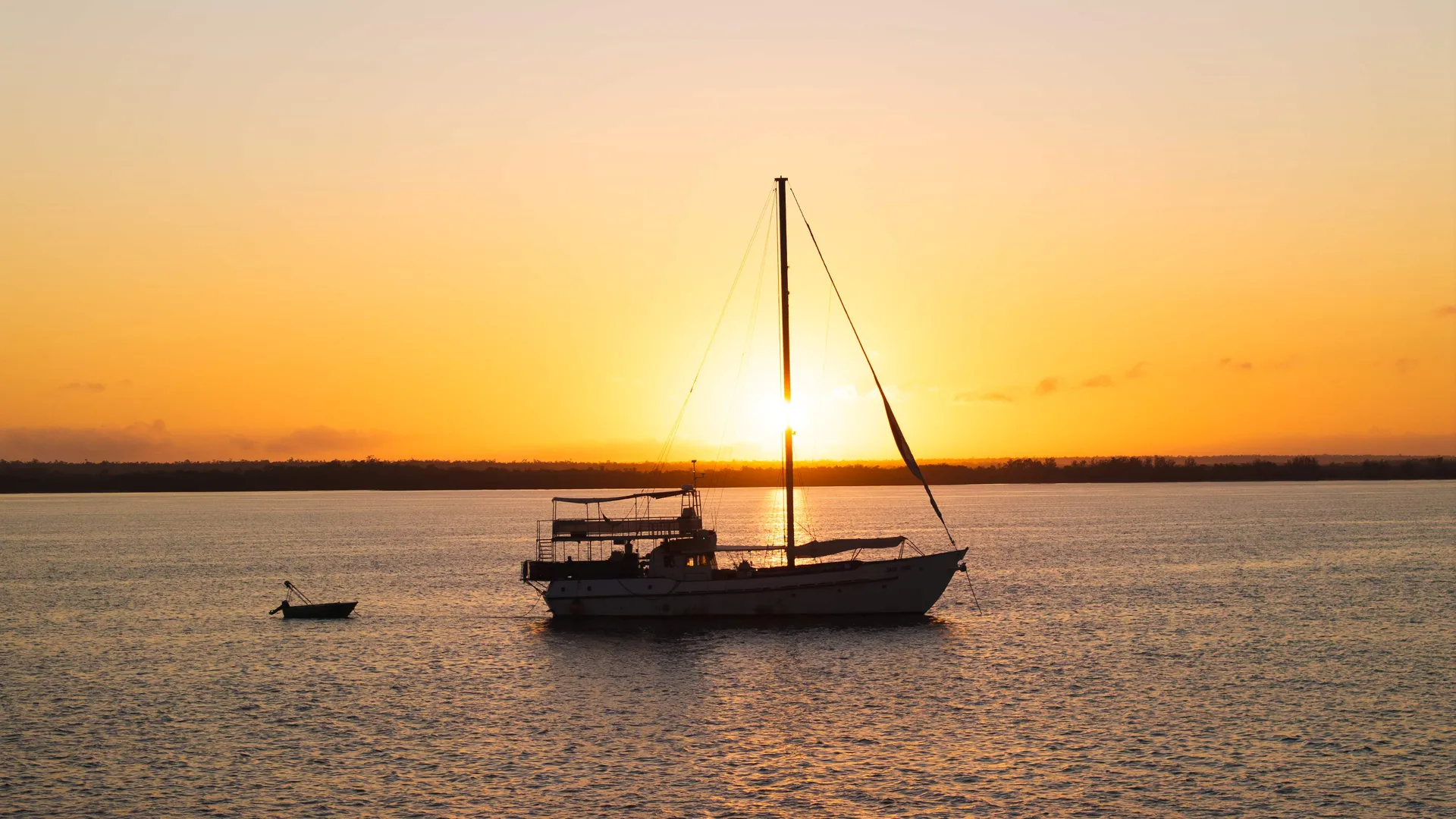 A sailboat anchored on calm water at sunset, silhouetted against a glowing golden-orange sky. The sun sits low on the horizon behind the boat's mast, casting shimmering reflections across the water.