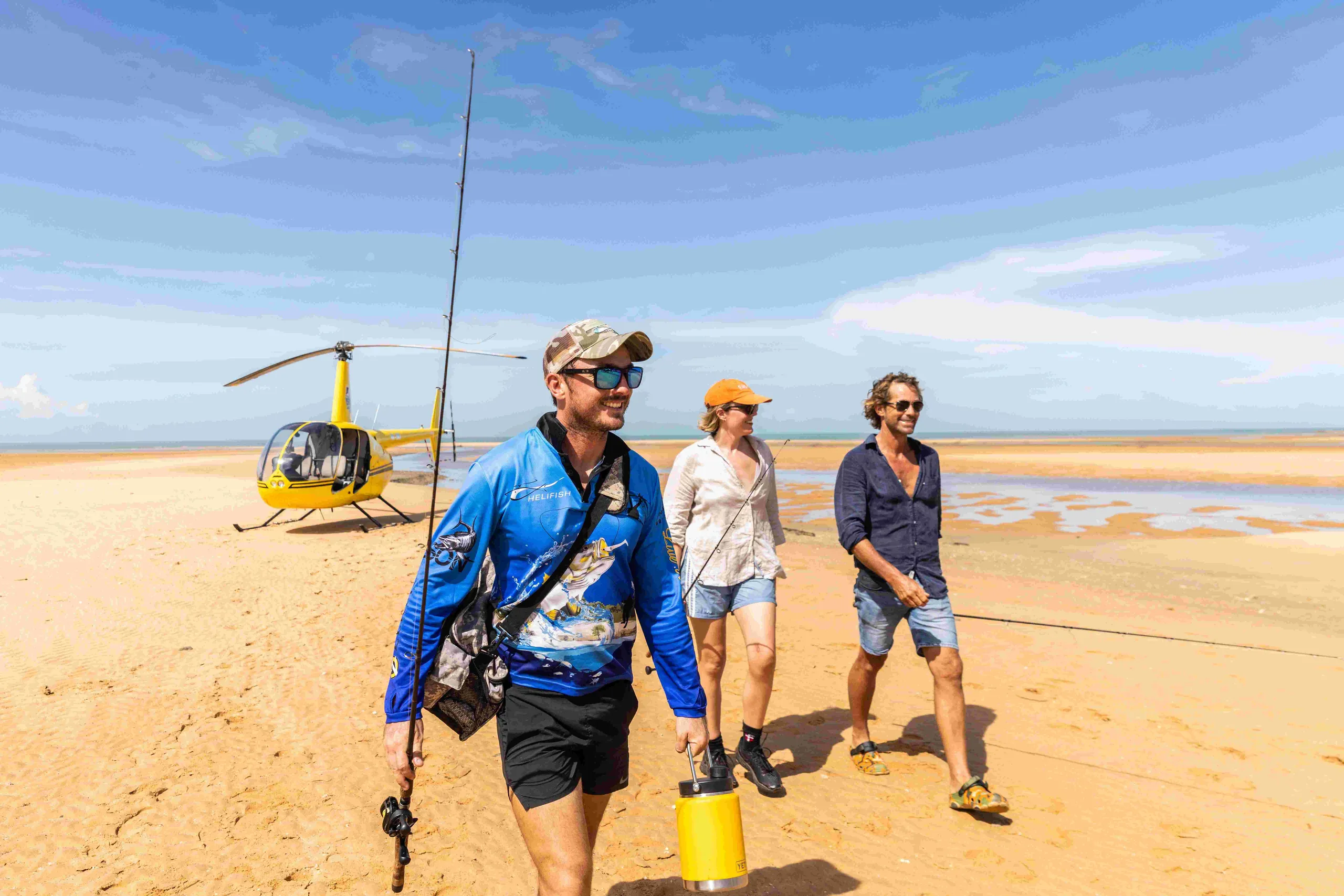 Three anglers carrying fishing gear walk across a remote beach beside a yellow helicopter on a coastal tidal flat.