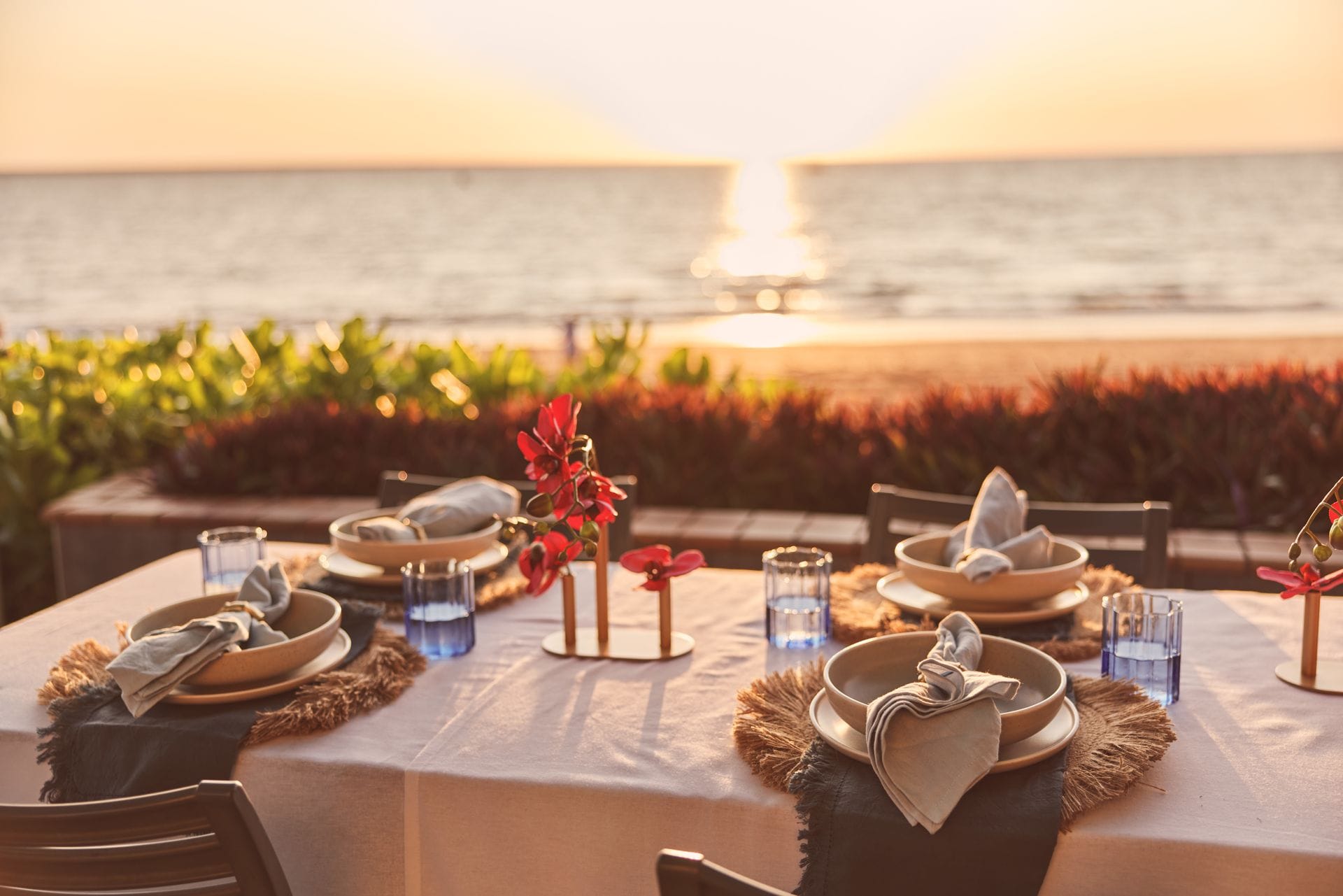 Elegant beachside dining setup with plates, napkins, and red floral centrepieces on a table, overlooking a sunset-lit ocean.